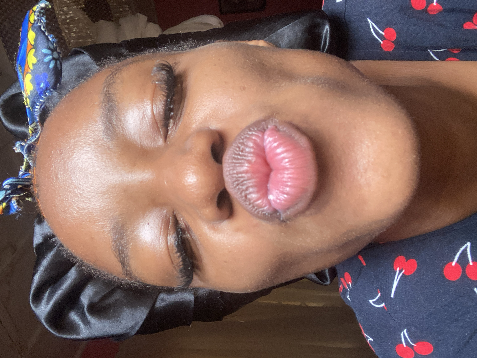 A close-up, frontal portrait features a person with dark brown skin, presumed female, in the city of Jos, Nigeria. The subject's eyes are closed, and the lips are puckered into a "kiss" shape, exhibiting a glossy, pink appearance. Elongated dark eyelashes are visible on both eyelids. The subject wears a black satin bonnet that covers the hair, and a small, colorful fabric headband with blue, yellow, and orange patterns is positioned atop the bonnet. The upper portion of a dark-colored garment, adorned with a repeating pattern of red cherries with white stems, is visible on the subject's chest and shoulders. The background is dimly lit and largely out of focus, indicating an indoor environment, with indistinct shapes and varying shades of dark brown, red, and off-white visible behind the subject. A faint reflective object, possibly a light fixture, is partially visible at the top right edge of the frame.