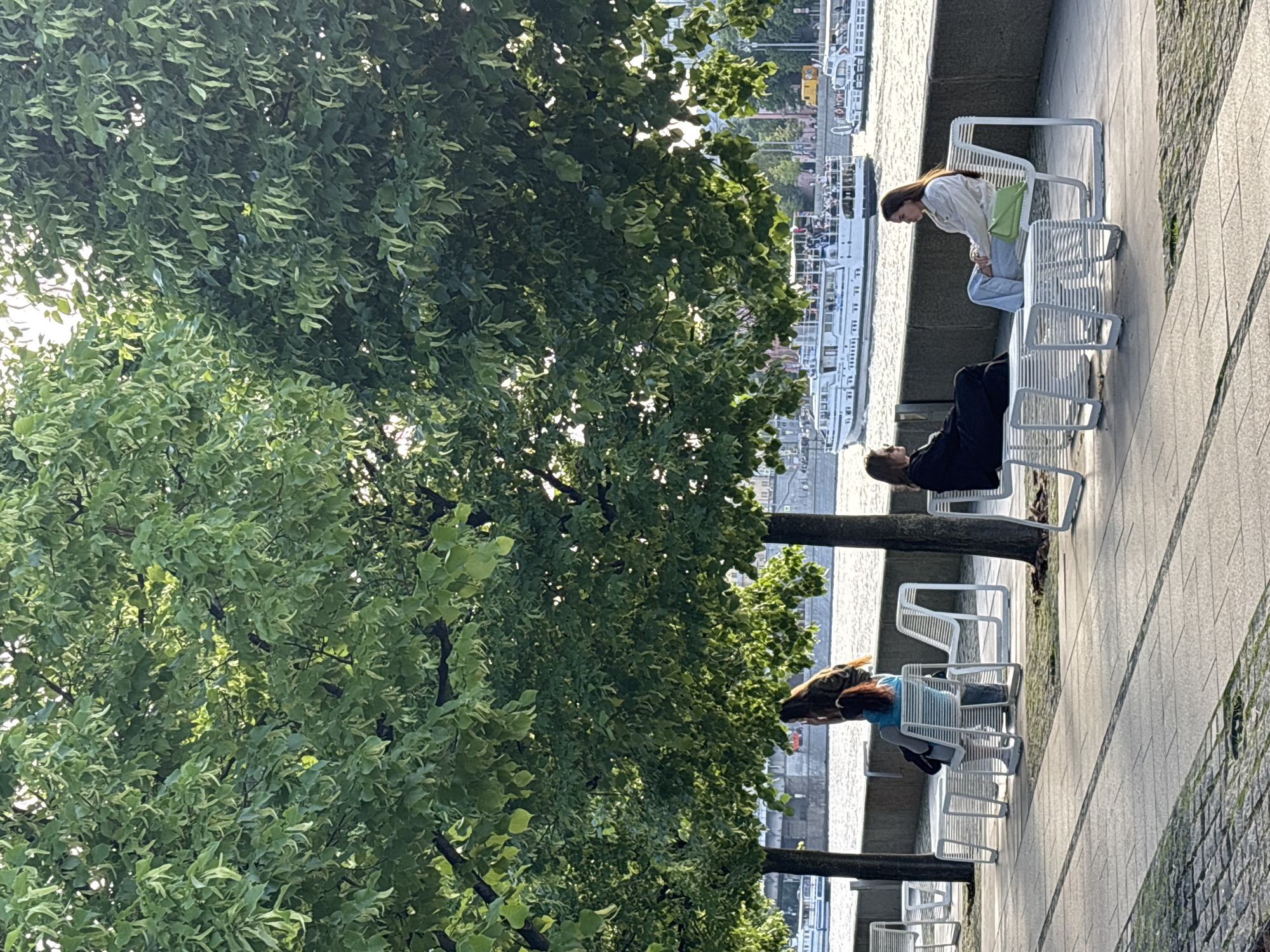 The image shows a waterfront scene in what appears to be an urban area, likely Moscow, Russia, based on the style of architecture and river boats visible in the background. Three people are sitting on modern, white, metal benches along a stone embankment or walkway. A large, leafy tree dominates the left side of the image, partially obscuring the view but also providing shade. The first person is standing over the bench, the second person is sitting upright, and the third person is sitting with their head back. In the background are several boats along a river. The scene appears to be taken during the day, with sunlight filtering through the trees.