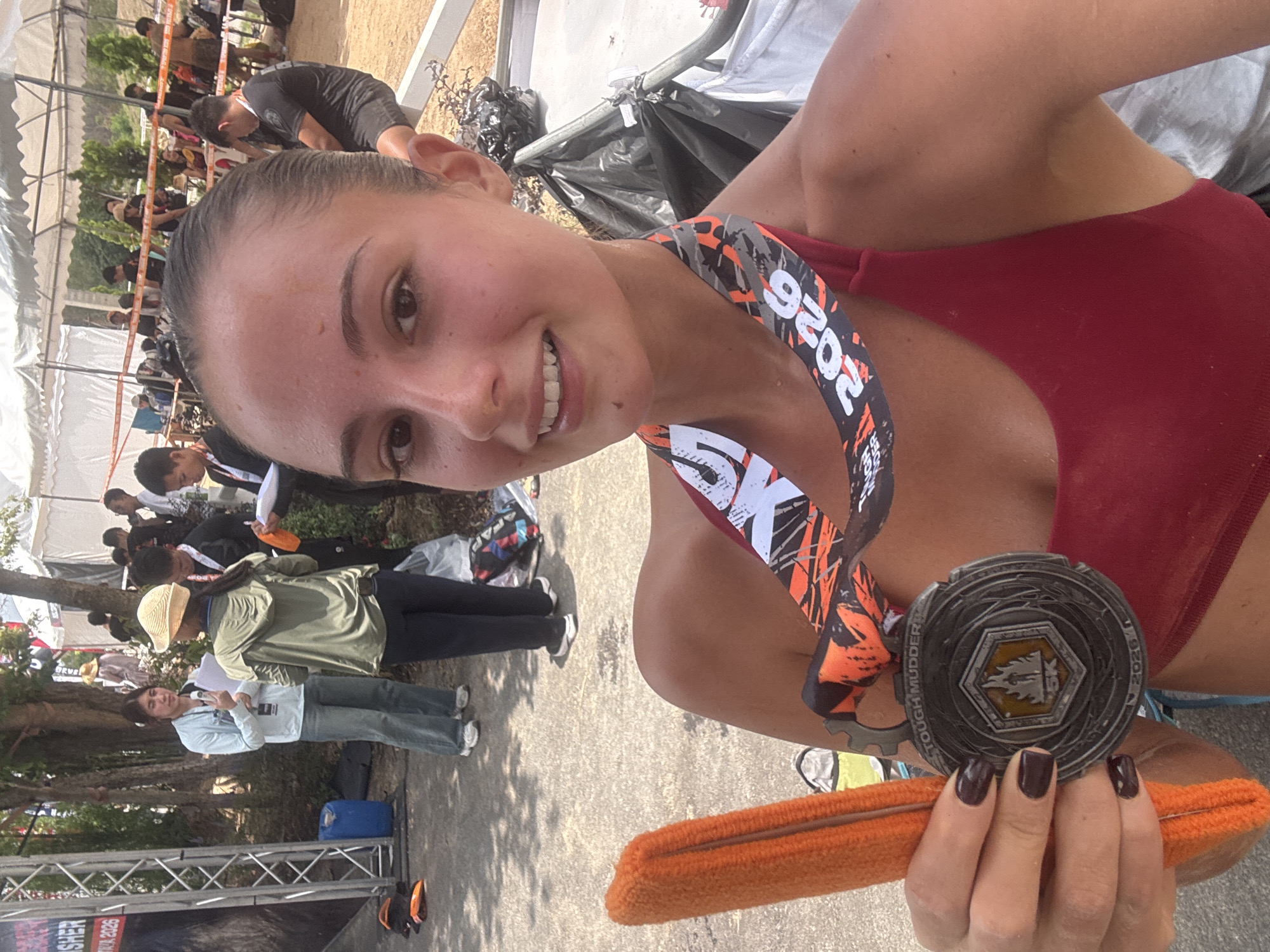 A selfie taken in บ้านสำนักตะแบก, Thailand, depicts a woman with light skin, dark hair pulled back, and wearing a red sports bra. She holds a "Tough Mudder 5K 2026" medal in her left hand, along with an orange headband. The medal hangs around her neck with a black, white, and orange patterned ribbon. The woman is smiling and looking at the camera.
The background shows an outdoor setting with people and structures. There are figures wearing various clothing, and a tent structure with an orange barrier is visible. Some trees and foliage can also be seen. The ground appears to be paved with concrete or asphalt.