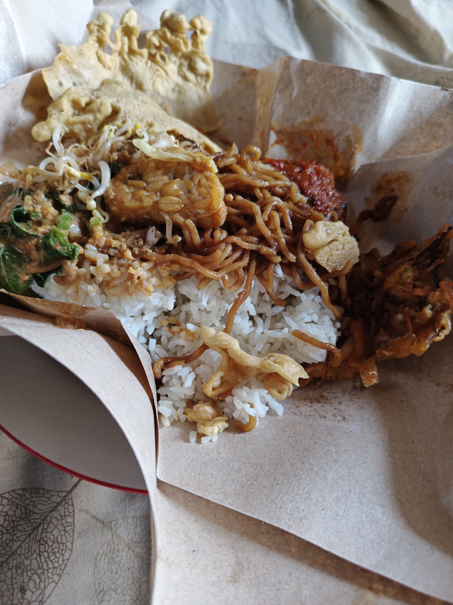 A close-up, overhead shot shows a plate of Indonesian food served on brown paper. The meal consists of white rice, topped with fried noodles, tempeh, bean sprouts, and a dollop of chili paste. Several fried crackers, one with an intricate, raised pattern, are also present. The food is arranged in a slightly messy, rustic presentation, suggesting it was takeout or a casual meal. The background is a light-colored, textured fabric, possibly a tablecloth. The image appears to be taken in the city of Rowokangkung, Indonesia.