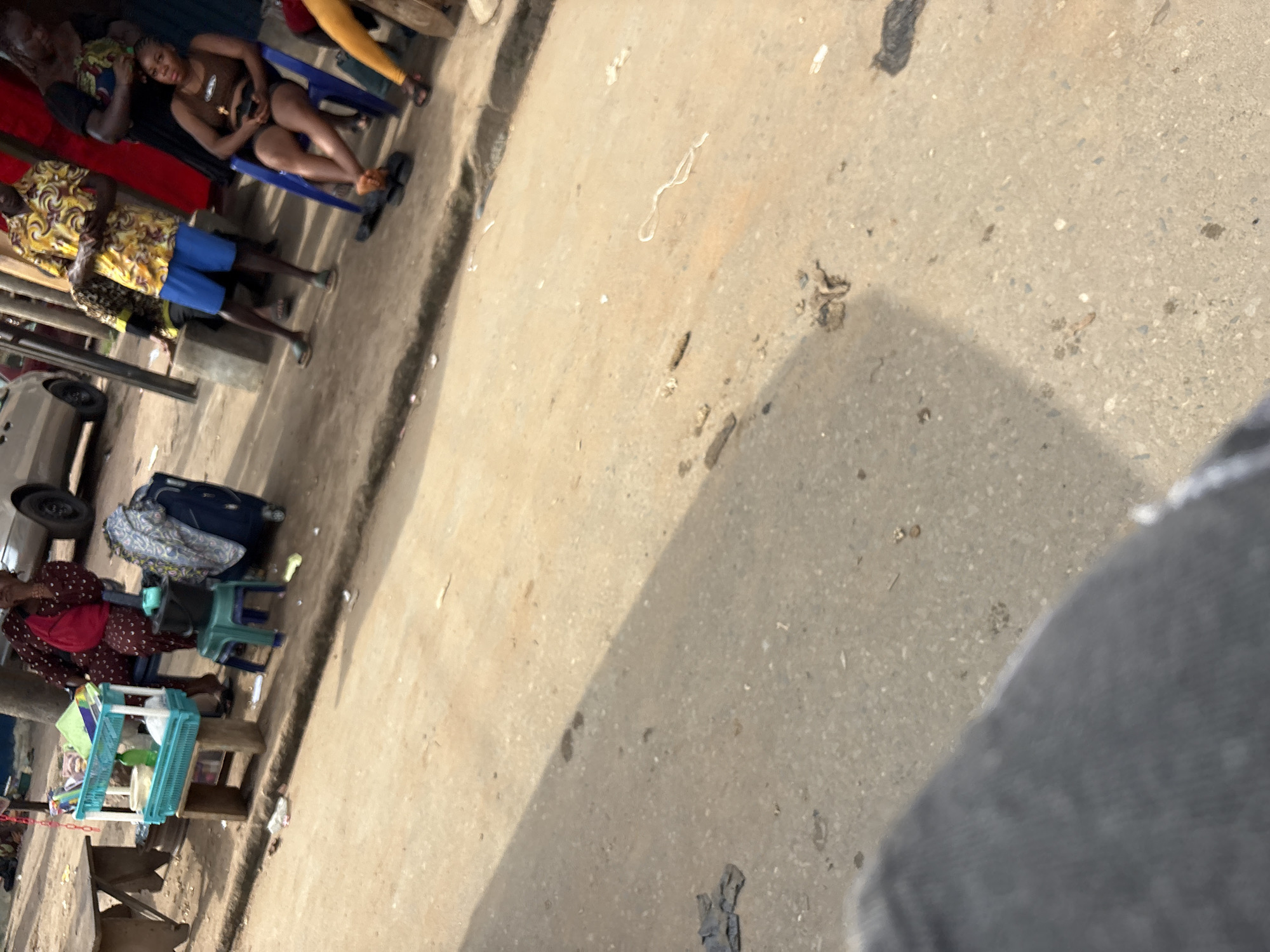 The image depicts a street scene in Onitsha, Nigeria, likely a market or roadside vendor area. In the foreground, a person's legs and lower torso are visible, clad in dark grey trousers or jeans, suggesting the photographer is standing or walking. The ground is paved with asphalt, exhibiting a rough texture and scattered debris, including some small, dark stains and a white string.
Further into the scene, a young woman is seated on a blue plastic chair, wearing a brown tank top and shorts. She is holding a dark object, possibly a phone, in her hands. To her left, a man in a bright yellow and black patterned shirt and blue shorts stands, also looking down at an object in his hands. Behind him, another person in a patterned garment is partially visible.
To the left of the seated woman, a woman in a dark red and white patterned dress is seated. She has a red bag slung over her shoulder. Next to her is a blue and white plastic stand holding various items, including a green bottle and white containers. A dark suitcase is visible behind the woman in the red dress. A small green plastic stool is placed near the blue stand. In the background, a portion of a silver car and other structures are visible, indicating an urban setting. The overall impression is of a busy, outdoor commercial environment.