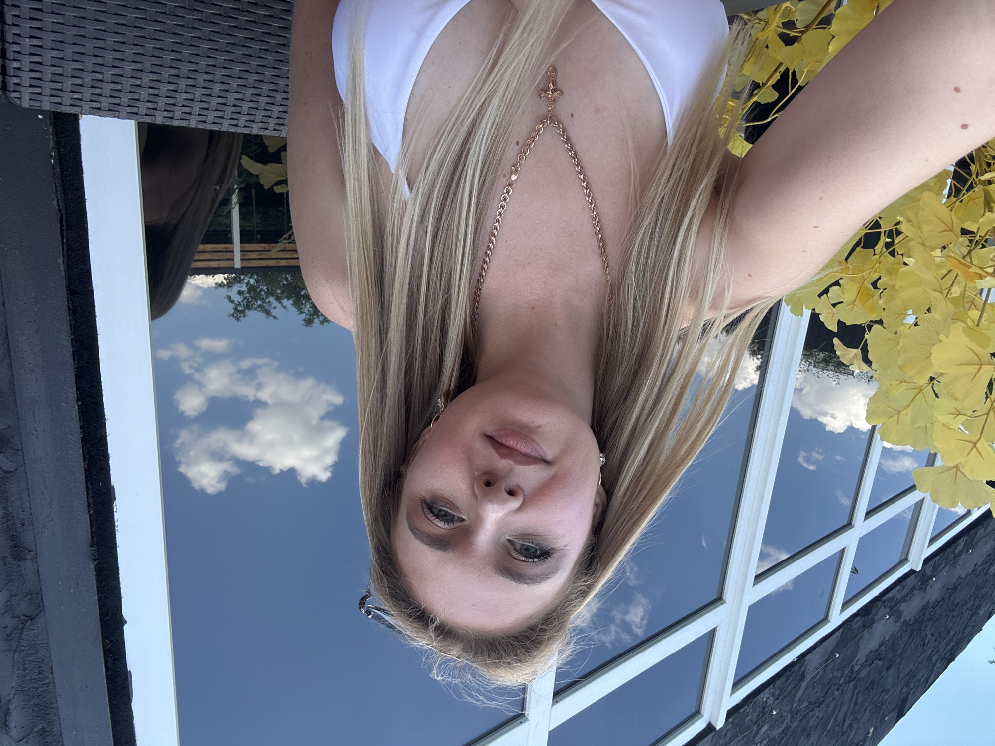 A blonde woman in a white bikini top and a gold cross necklace is shown in a selfie. She is sitting outside, with a large window reflecting the blue sky and clouds behind her. Yellow foliage is visible to her left. The scene appears to be in a relaxed, outdoor setting, possibly a cafe or restaurant.
