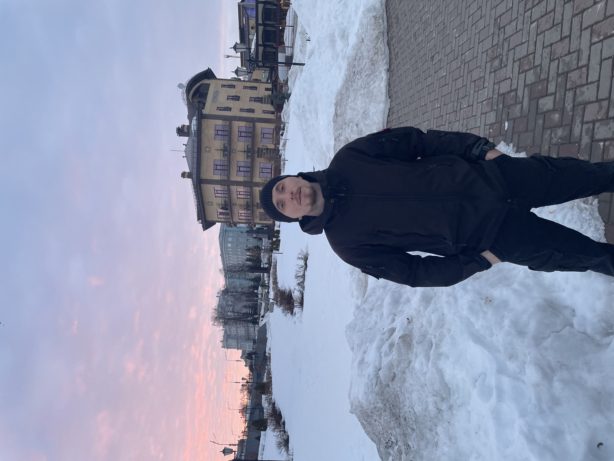 A male individual is depicted standing outdoors in what appears to be a city park or public space in Sarapul, Russia, during twilight conditions (either dawn or dusk). The man is positioned centrally, facing directly towards the camera. He has short facial hair, a light beard/stubble, and is wearing a black knitted beanie. His attire consists of a dark-colored, possibly black, hooded jacket with a zipper, featuring a red and white patch on the upper left arm, and dark pants, likely cargo-style, with his hands placed in his pockets. The ground beneath him is paved with reddish-brown interlocking bricks, while the surrounding landscape is extensively covered in snow, forming uneven mounds and drifts, some appearing soiled.
In the background, a multi-tiered urban environment is visible. To the man's left, an elevated terrace features a dark metal railing, bare trees, and streetlights, with a large, light-colored multi-story building (possibly grey or light blue) visible further back on a higher elevation. To the man's right, a distinctive yellowish-brown brick building with dark window frames, multiple balconies, and a curved architectural design is prominent. The word "ОТЕЛЬ" (HOTEL) is visible vertically on its facade. The sky above presents a gradient of colors, transitioning from pink and orange near the horizon to a lighter blue-purple overhead, with scattered clouds.