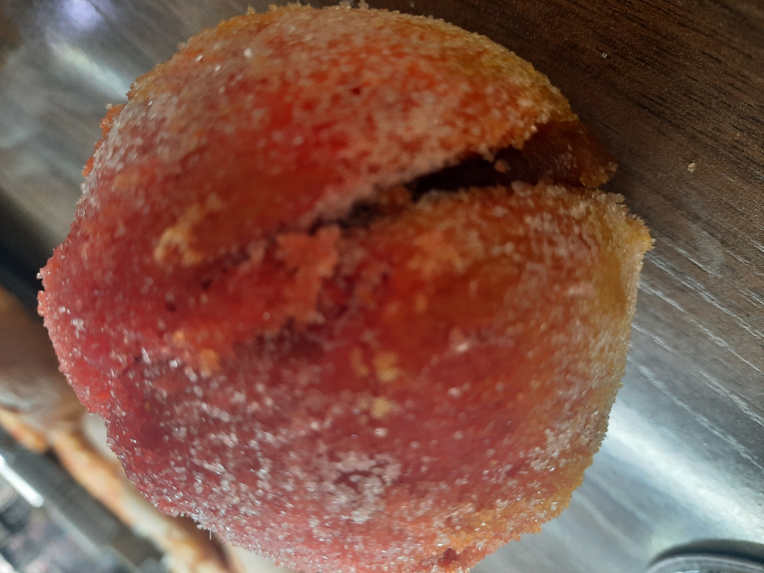 A close-up, eye-level photograph captures a single, round pastry or confectionery item. The item is vibrant reddish-orange and heavily coated with glistening white sugar crystals. A distinct split or crack is visible on its top surface, revealing a darker, rich brown filling or interior. The sugar coating appears granular and prominent, sparkling under the soft lighting. The pastry rests on a surface with a visible wood-grain pattern, possibly a table or counter. The background is blurred and indistinct, ensuring the focus remains entirely on the food item. No people, activities, or discernible text are present in the image.