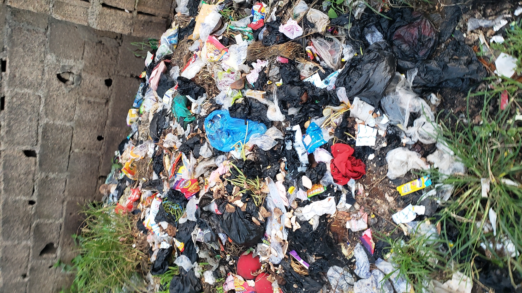 The image depicts a large pile of garbage against a grey brick wall. The garbage consists of various plastic bags, wrappers, food scraps, and other debris. Some green grass is growing around and through the trash. A bright blue plastic bag is visible in the center of the pile. The scene appears to be an outdoor area, possibly an alleyway or an undeveloped lot. There are no people or animals visible in the image. The lighting suggests it is daytime. The context of Jalingo, Nigeria, implies this is an urban or semi-urban environment where waste management may be a challenge.
