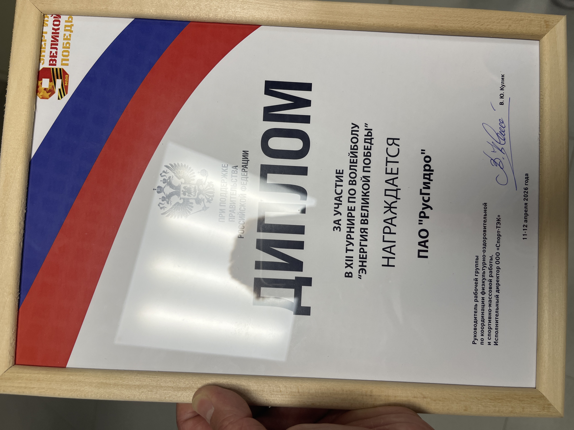 A framed diploma is presented. The diploma, with text in Russian, features the colors of the Russian flag along the left border and a stylized graphic commemorating Victory Day in the upper left. The certificate states "Diploma" and indicates participation in the 12th volleyball tournament "Energy of Great Victory". It awards PAO "RusHydro". The diploma bears the name "V. Kulik" and a signature. It is dated April 11-12, 2026. The diploma mentions support from the government of the Russian Federation.