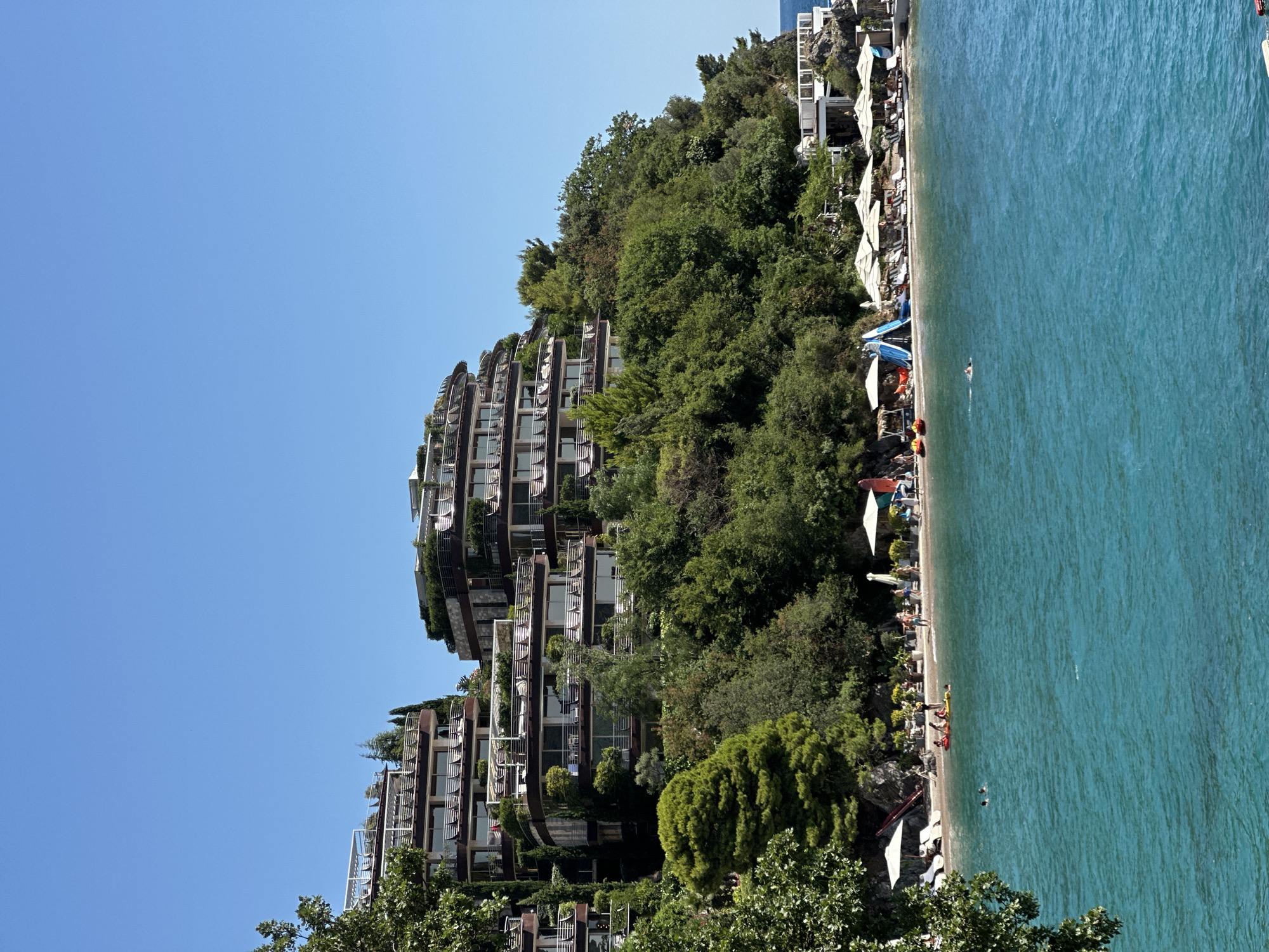 This is a landscape photograph of a resort located on a hillside overlooking the Adriatic Sea in Budva, Montenegro. The resort consists of multiple terraced buildings with curved facades and balconies, partially covered in vegetation. A beach with lounge chairs and umbrellas is situated at the base of the hill, with several people visible on the sand and in the water. The sea is a vibrant turquoise color, and the sky is clear blue. In the foreground, a tree branch partially obstructs the view on the left side of the image.