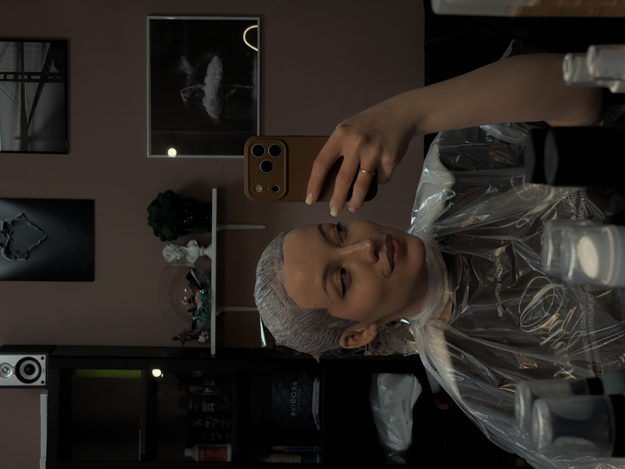 A person with a head covered in hair dye, wearing a clear plastic cape, is taking a selfie with a brown smartphone. The person is wearing a ring on their left ring finger. The background includes a wall with black and white photographs, a shelf with decorative items, and a black cabinet containing various products. The location is unknown.