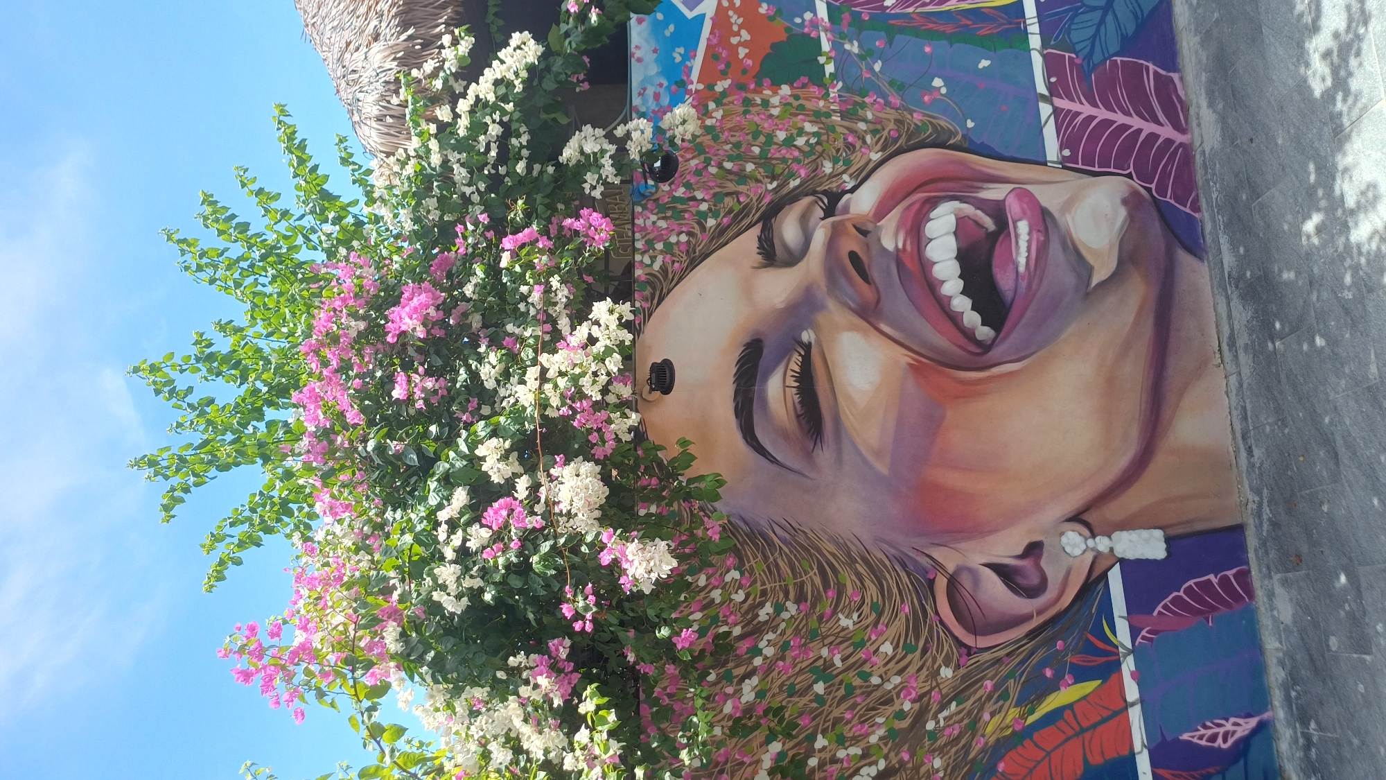 A close-up, eye-level, outdoor medium shot captures a colorful mural of a woman's face on a wall, with vibrant bougainvillea flowers and green foliage arching over her head. The mural is located in Da Nang, Vietnam, likely a street art district or a public space adorned with art. The woman in the mural is depicted with her eyes closed, mouth open in laughter or exhilaration, revealing white teeth and a pink tongue. Her hair is stylized with painted flowers and organic patterns. She wears a long, white dangle earring. The background wall features additional graphic elements, including large, stylized leaves in shades of purple, pink, and blue. To the upper right of the mural, a portion of a thatched roof is visible, suggesting a tropical or beachside setting. The sky is bright blue with wispy white clouds. The ground in front of the mural is paved with grey cobblestones, some of which are shaded by dappled sunlight.