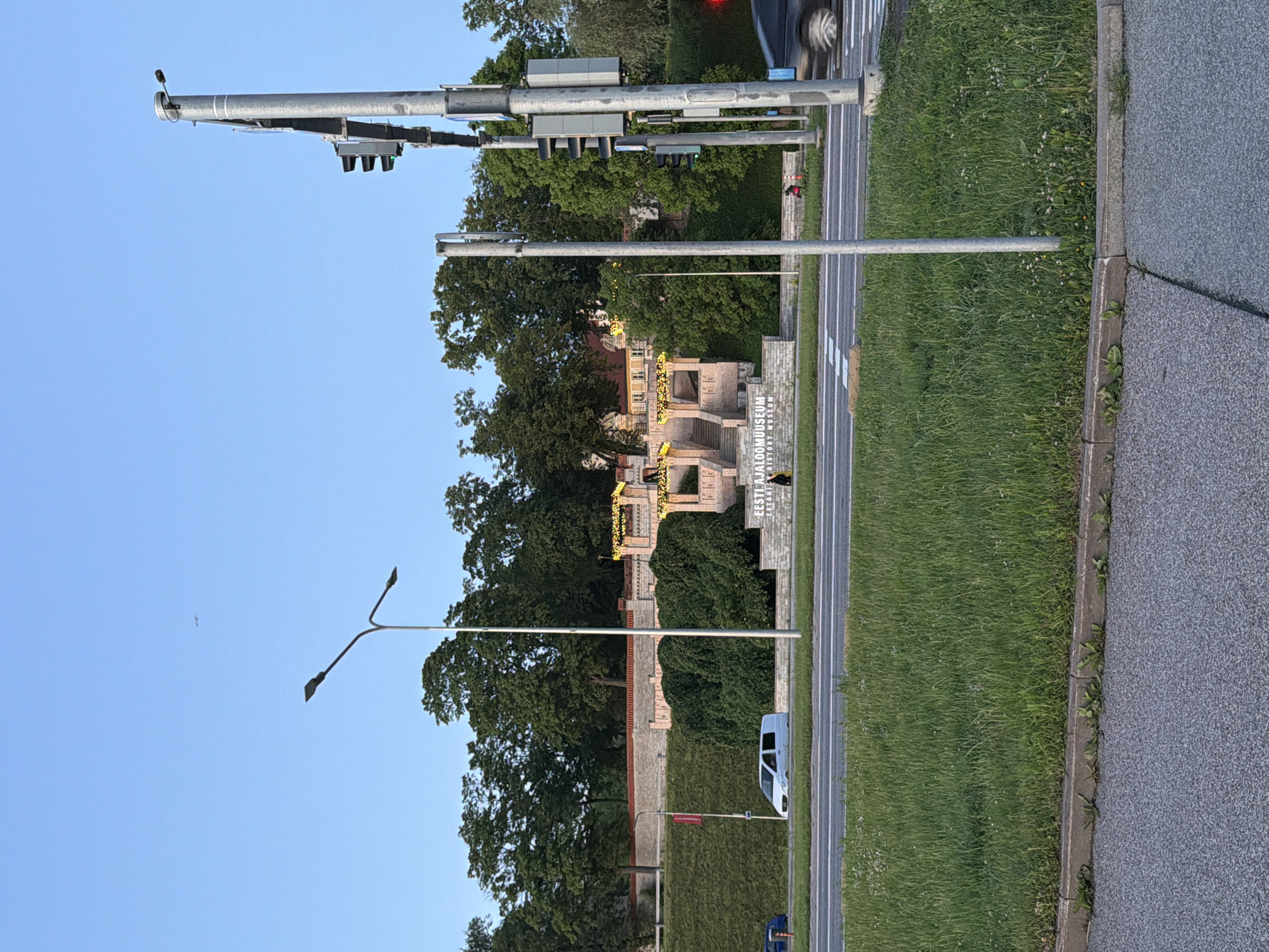 The image shows the Estonian History Museum (Eesti Ajaloomuuseum) in Tallinn, Estonia, on a clear day with a bright blue sky. In the foreground, a paved road runs horizontally across the image, with a grassy median separating it from another lane of traffic. A white van is parked on the left side of the road, and a portion of a dark-colored car is visible on the right, indicating movement.
Behind the road and median, a large stone building with decorative yellow lights and stairs leading up to it is visible. The text "EESTI AJALOO MUUSEUM" and "HISTORY MUSEUM" are inscribed on a stone wall in front of the building. Lush green trees and bushes surround the museum, with a red-tiled wall and a grassy embankment visible behind the museum.
Two streetlights are prominent in the image; one on the left with two lamps, and a taller one on the right with multiple traffic lights and a lamp at the top. Several poles with electrical equipment are also present near the traffic lights. A few people are scattered in the background, one walking near the museum and another on a red bicycle further to the right. The overall scene depicts an urban environment with historical architecture and a touch of nature.