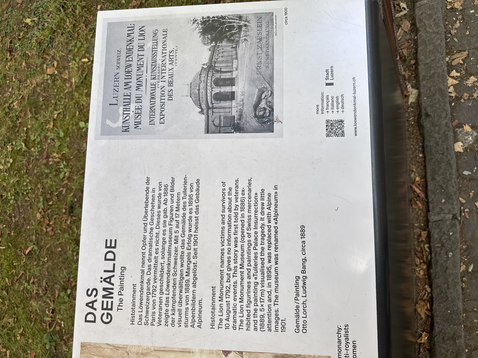 The image shows an outdoor informational sign with text in German and English, alongside a black and white illustration. The sign is positioned at ground level, with a grassy area and fallen leaves visible in the foreground.
The left side of the sign features a large heading "DAS GEMÄLDE" (The Painting) in German, followed by the English translation. Below this, there are two sections of text. The first section, titled "Histotainment," describes a painting that depicts the victims and survivors of August 10, 1792, and its exhibition history, including its placement in the Lion Monument Museum and eventual renaming to "Alpineum." The second section provides details about the painting itself, listing "Gemälde/Painting" and crediting "Otto Lorch, Ludwig Bang, circa 1889." At the bottom left, there are partial words "monarchy" and "ti-royalists" and "omen".
The right side of the sign displays a stylized illustration of a building with a dome, identified as "KUNSTHALLE AM LOEWENDENKMAL; MUSÉE DU MONUMENT DU LION" in German and French, indicating it is an art hall at the Lion Monument. Below this, it advertises an "INTERNATIONALE KUNSTAUSSTELLUNG (VERKAUF.) / EXPOSITION INTERNATIONALE DES BEAUX ARTS. (VENTE.)", which translates to "International Art Exhibition (For Sale) / International Exhibition of Fine Arts (Sale)". The illustration itself includes text that reads "ERNST ZAESLEIN KUNSTHANDLUNG.", which translates to "Ernst Zaeslein Art Dealership". The illustration is dated "circa 1900".
At the bottom of the sign, there is a QR code and text providing language options ("français", "italiano", "english", "deutsch") and a website address: "www.loewendenkmal-luzern.ch". The town of Lucerne, Switzerland is mentioned in the text "Luzern Schweiz" and on the website. The overall context suggests the sign is located near the Lion Monument in Lucerne, Switzerland, providing information about a painting and an art exhibition.
