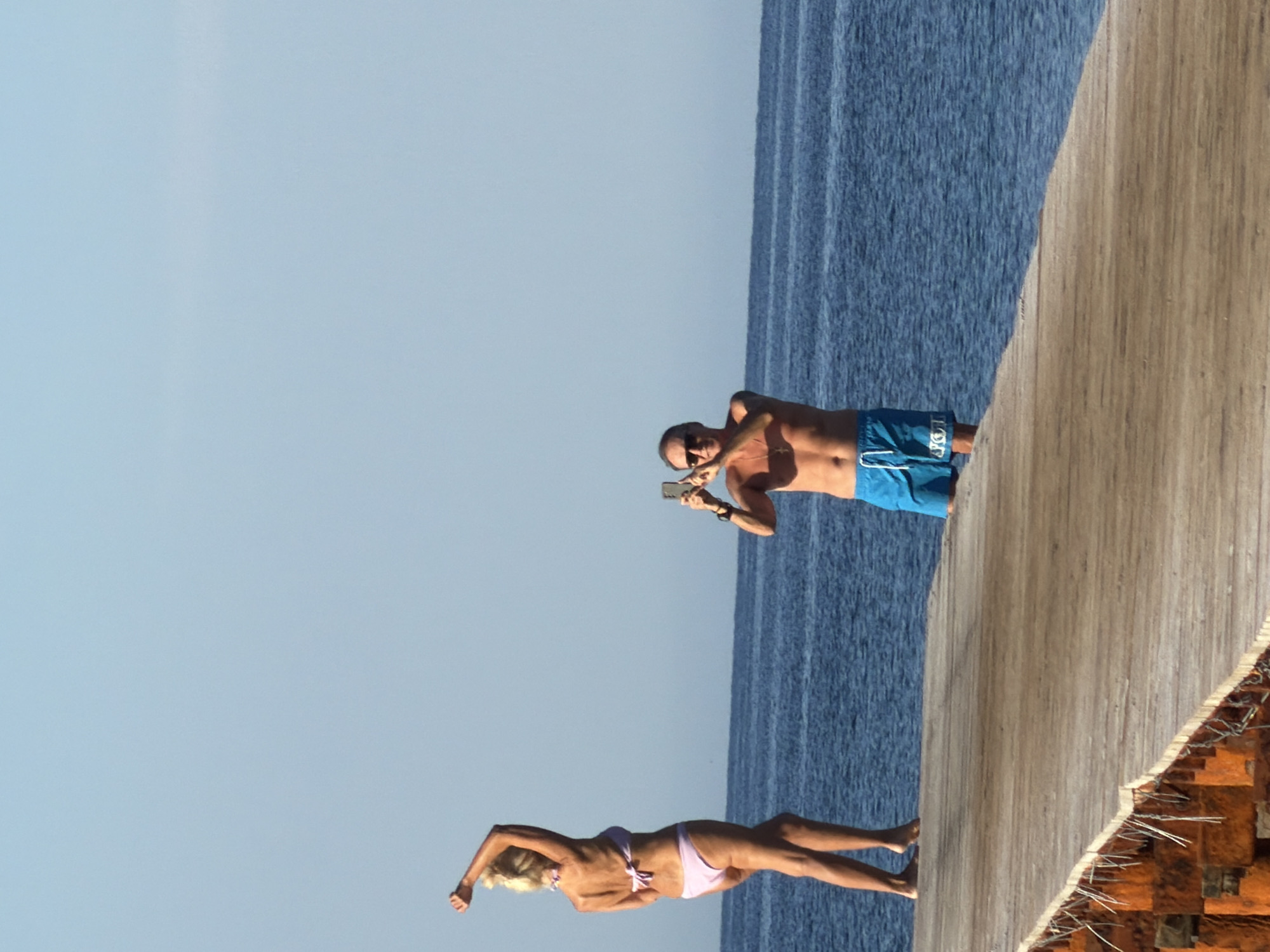 In Budva, Montenegro, on a wooden pier, a woman in a light purple bikini poses with her arms raised. To her right, a man in blue swim trunks holds up a smartphone, appearing to take a photograph. The scene is set against the backdrop of a clear blue sky and the calm, blue sea.