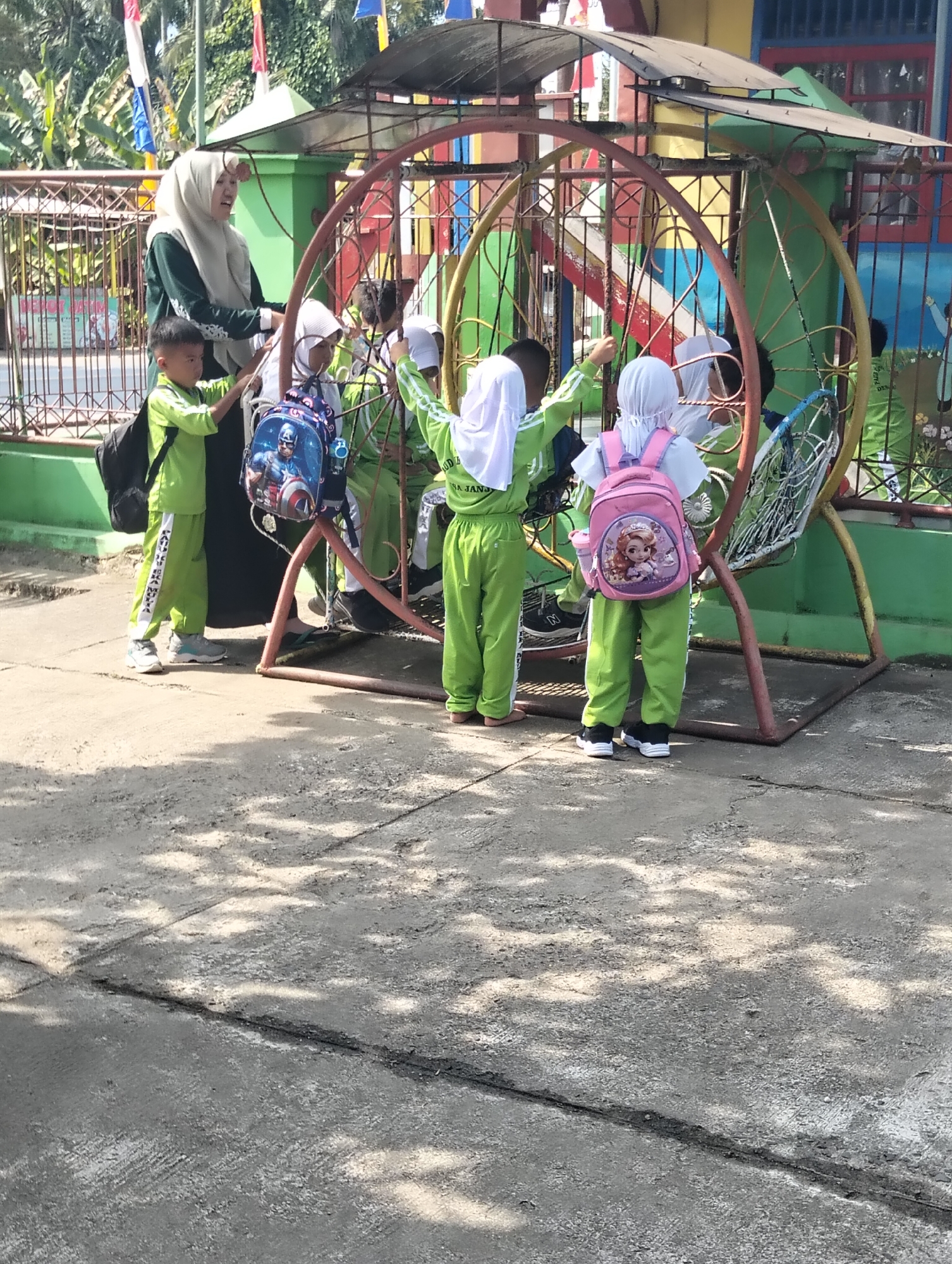 The image depicts a group of children in green school uniforms and headscarves gathered around a large, circular swing set in what appears to be a schoolyard. A teacher, wearing a dark green hijab and long dress, stands among them. Several children are wearing backpacks, including one with a Captain America design and another with a princess graphic. The swing set has a colorful, weathered appearance, with red, yellow, and green accents. In the background, there is a green fence, some foliage, and a building with colorful walls, suggesting an educational facility. The ground is paved with concrete, showing some cracks and shadows from overhead trees. The scene appears to be in an outdoor, daytime setting. The image is consistent with a playground or school area in Janju, Indonesia, given the architectural style and vegetation.