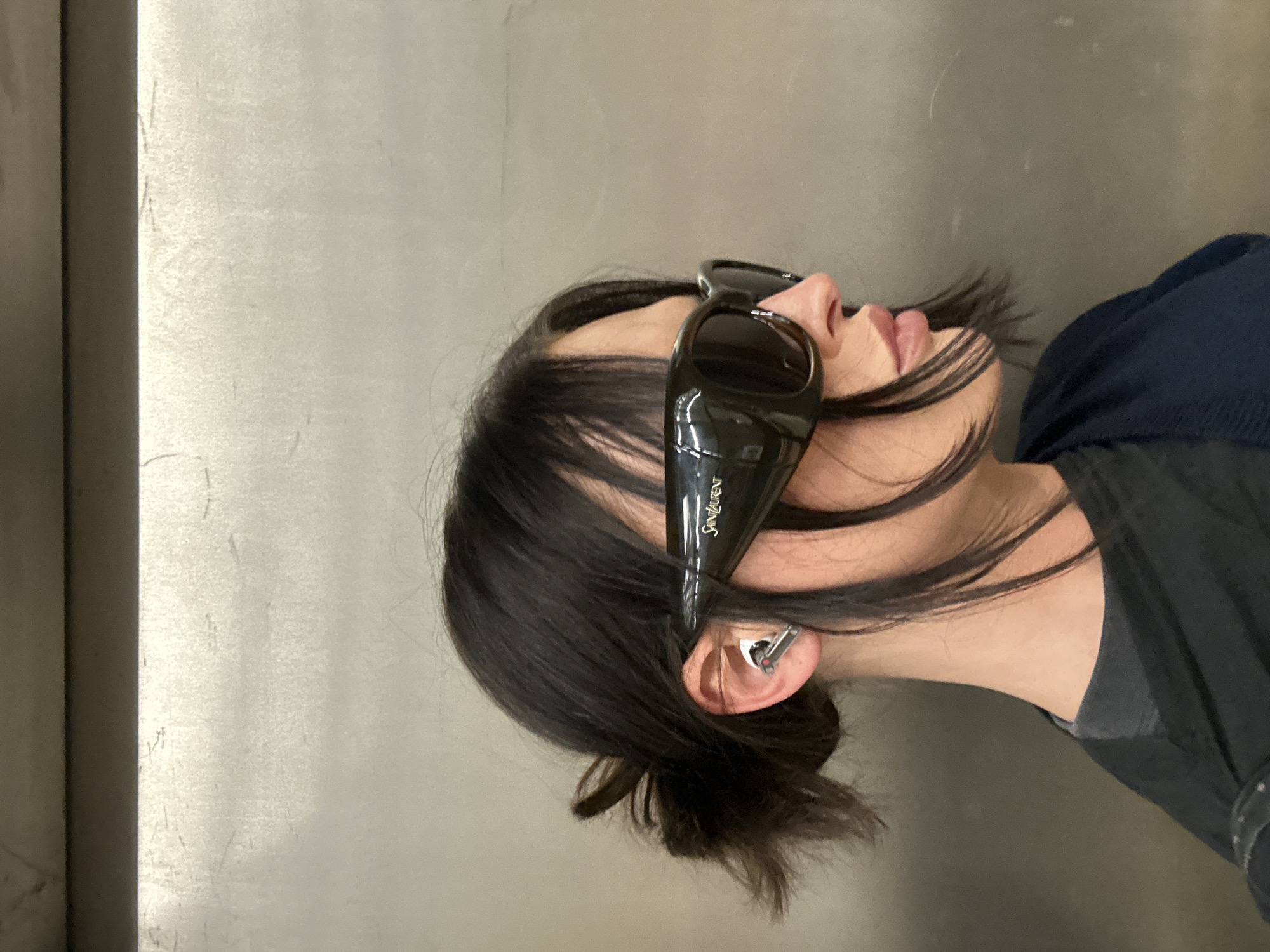 A subject, viewed in profile facing right, is positioned against a light grey, horizontally textured metallic wall with a dark horizontal structural element at the top. This environment is consistent with a public transport interior, such as a London Underground station or an elevator, within the city of London, United Kingdom. The subject exhibits dark hair styled in a low bun with loose facial strands, and wears oversized dark wrap-around sunglasses marked 'Saint Laurent' on the right temple. A silver-toned wireless earbud is present in the left ear. Clothing consists of a dark t-shirt beneath a navy blue cardigan draped over the shoulders.
