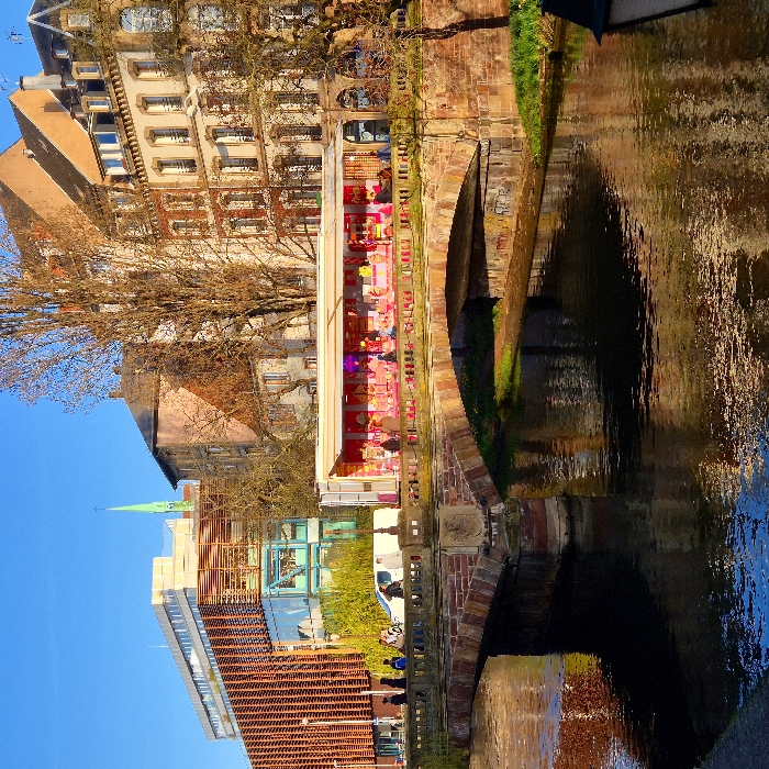 The image captures an urban waterscape in Strasbourg, France, specifically at the Pont Sainte-Madeleine crossing a canal, likely the Canal du Faux-Rempart. The scene is illuminated by bright sunlight under a clear blue sky, casting distinct shadows suggestive of early morning or late afternoon.
A multi-arch bridge constructed of reddish-brown stone masonry spans the canal. A crest or coat of arms is embossed on the central pier of the bridge. On the bridge deck, a prominent elongated white structure with a red awning functions as a temporary market stall or vendor, brightly lit internally and displaying various items, possibly food or merchandise. Multiple indistinct figures are present within and around this stall, engaging in browsing or service. To the left on the bridge, a white van is parked, and a group of approximately five to seven pedestrians are walking. A dark lamppost is visible on the bridge parapet.
On the left bank of the canal, a modern building, identifiable as the Médiathèque André Malraux, features a facade of vertical wooden louvers and large glass sections. Beyond this, further in the background, a slender green spire, consistent with that of Cathédrale Notre-Dame de Strasbourg, rises above the cityscape. On the right bank, a row of traditional multi-story buildings with warm-toned facades, numerous windows, and pitched roofs are visible. Bare deciduous trees, devoid of foliage, are interspersed throughout the background and on the right side of the frame. The canal water below is calm, reflecting the sky and the surrounding structures.