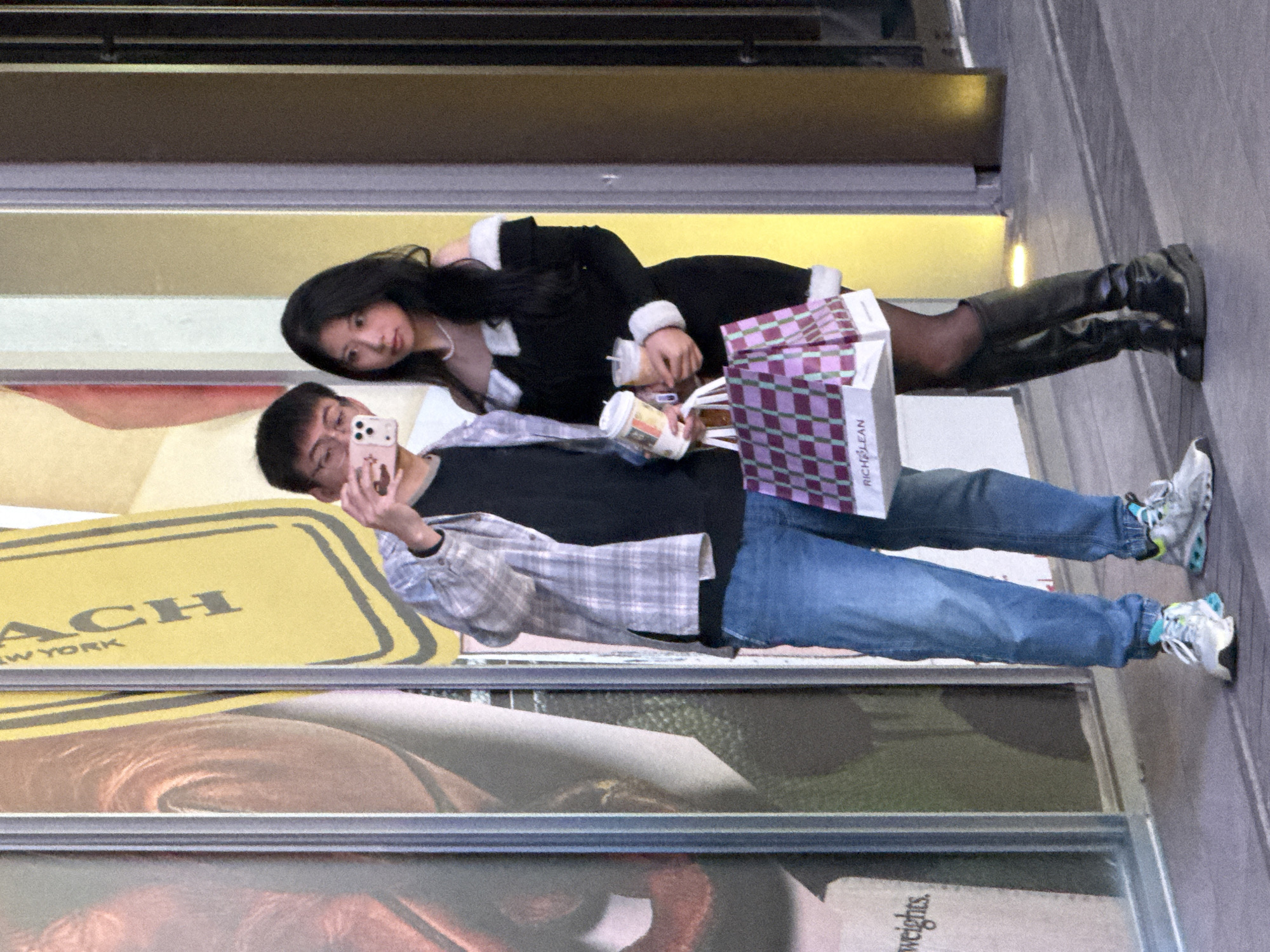 Two East Asian individuals, a male and a female, stand on a paved surface in front of a commercial building with large display windows in Liangjiang, China. The male, wearing a light plaid long-sleeve shirt over a dark t-shirt, blue jeans, and athletic shoes, holds a light pink smartphone in his right hand, extended outwards as if taking a photograph. He also holds one white paper beverage cup in his left hand. The female, dressed in a black off-the-shoulder short dress with white fur trim, black opaque tights, and black knee-high boots, holds two white paper beverage cups in her left hand and a purple and pink checkered shopping bag with "RICH&LEAN" branding in her right hand, which the male also grasps. The building's large display windows behind them feature vertical advertisements, including a yellow panel displaying "COACH NEW YORK" text. The scene is well-lit, indicative of an urban commercial environment.