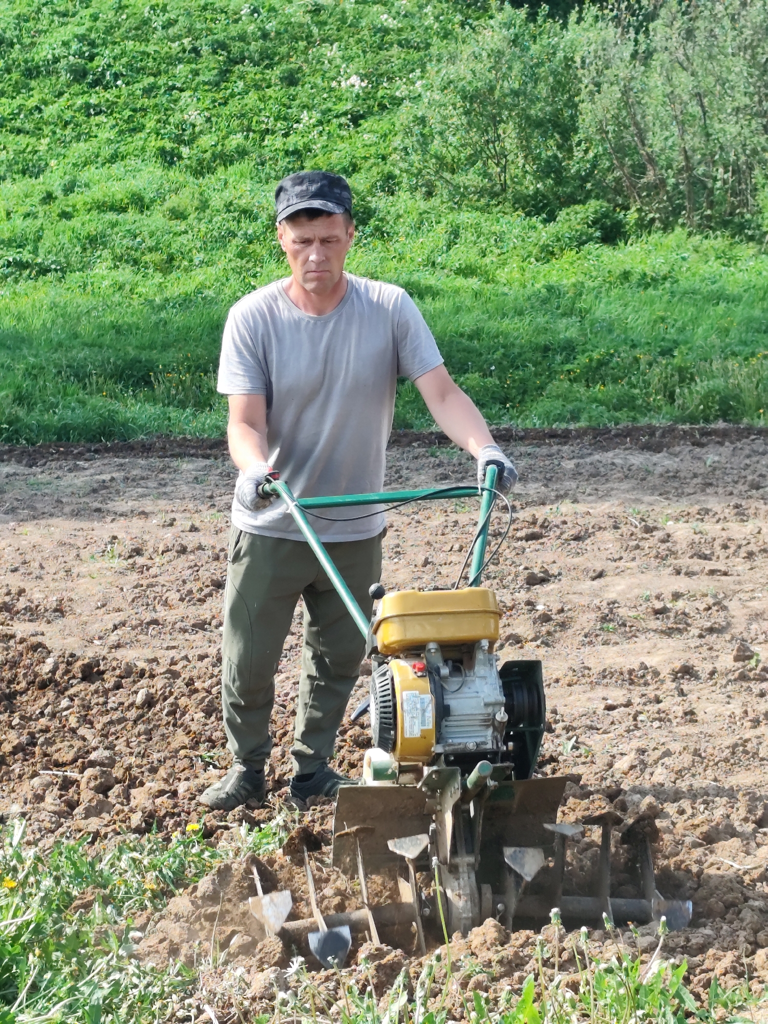 A man is operating a rototiller in a plot of land. He wears a gray t-shirt, dark cap, olive-colored pants, gloves, and dark shoes. The rototiller is predominantly yellow and black with green handles. The ground is tilled brown soil, with some grass and weeds visible. A grassy hillside is in the background. The location is Фоки, Russia.