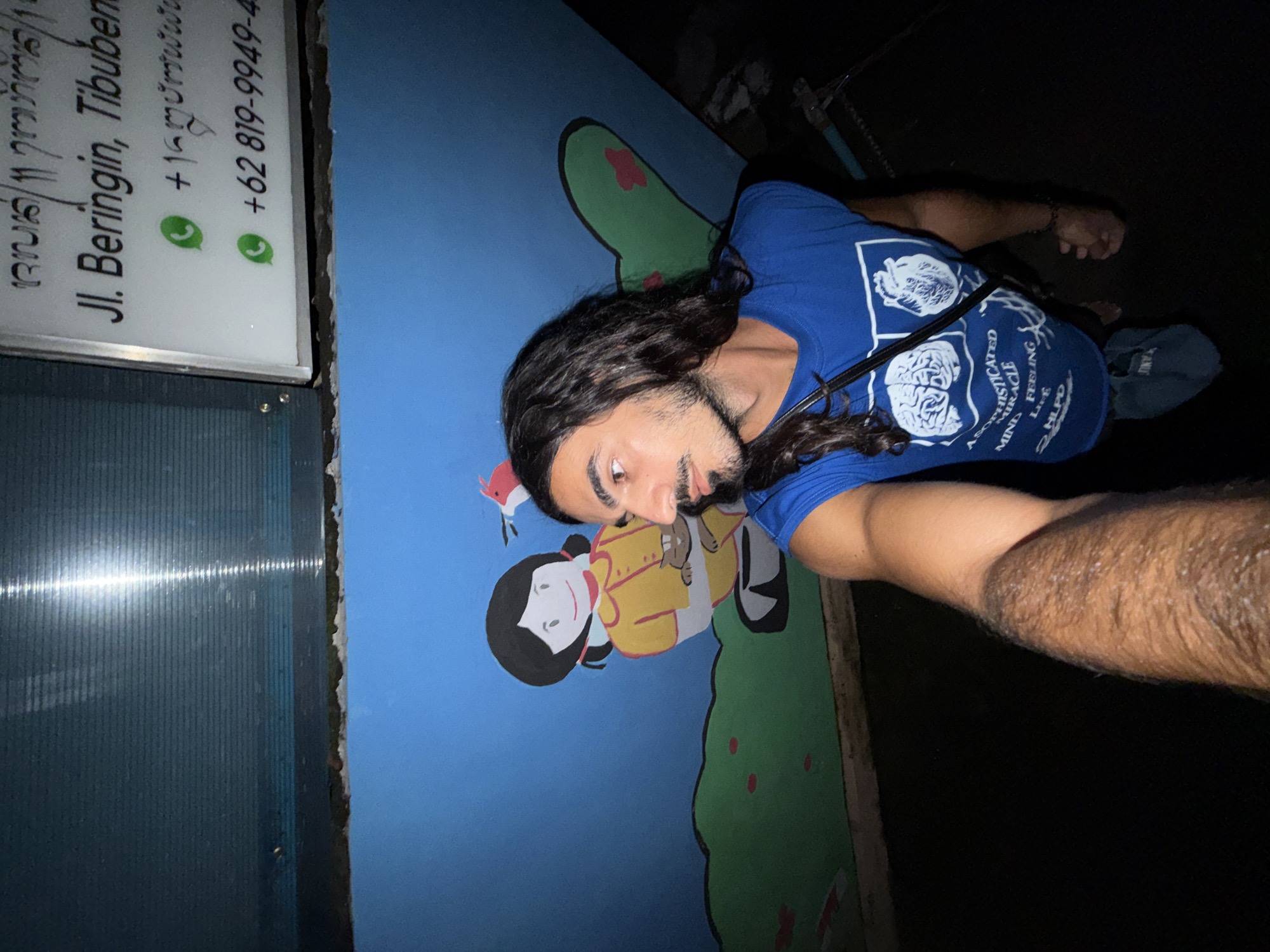 A man with long, curly hair and a beard is taking a selfie next to a mural depicting a Japanese cartoon character with a small bird perched above its head. The mural is painted on a blue wall with green patches and red accents. To the left of the mural is a sign that reads "Jl. Beringin, Tibuben" and includes a phone number. The man is wearing a blue t-shirt with white graphics and has a bag slung over his shoulder. The location is North Kuta, Indonesia.