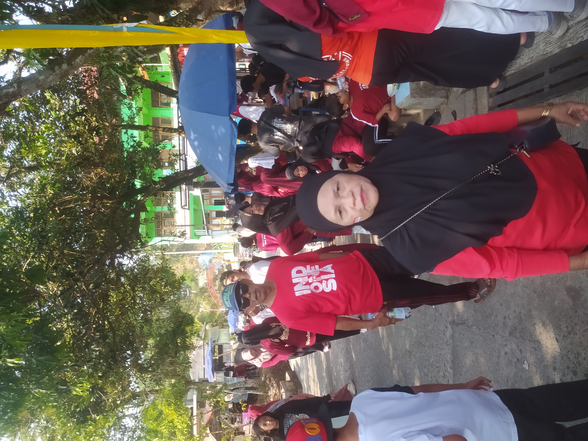 A crowd of people are gathered outdoors in Lopait, Indonesia, under a bright, sunny sky. The scene appears to be some sort of event or gathering. Many individuals are wearing red shirts, some featuring the word "INDONESIA" in white lettering. A man in the foreground wears a red shirt, sunglasses, and a blue cap, holding a water bottle. A woman in a black hijab and red shirt is also prominently featured. In the background, there are trees, a green building, and tents. The ground is paved, and there are some wooden benches. The atmosphere suggests a community event or celebration.