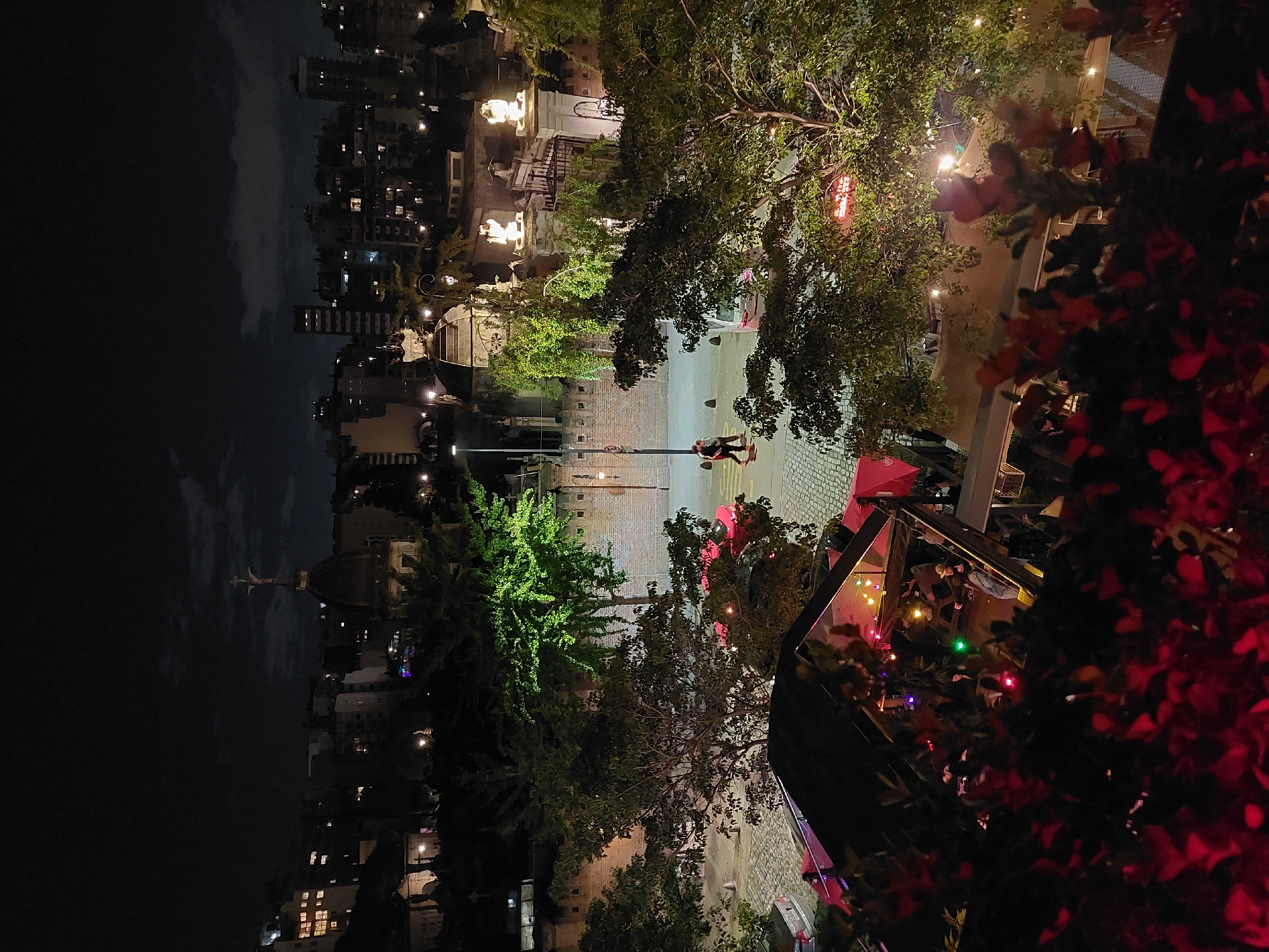 At night, the image depicts a street scene in Buenos Aires, Argentina. In the distance, illuminated buildings form a cityscape under a dark sky. The street itself is paved with cobblestones or bricks. Trees line the street and are interspersed with street lights. A person is visible near a traffic light. A building or structure, possibly a restaurant or cafe, is visible on the right side of the image, with red foliage in the foreground. The structure appears to have an open roof or terrace. String lights illuminate the area, suggesting a festive atmosphere.