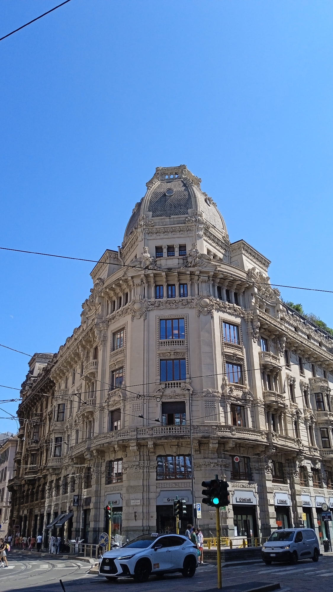 This is a low-angle, full shot of the historic Palazzo Castiglioni in Milan, Italy, on a clear, sunny day. The Art Nouveau building, known for its dome and ornate facade, is the dominant feature of the image. Electric wires stretch across the bright blue sky.
In the foreground, a street intersection is visible with a white Lexus NX 350h SUV stopped at a traffic light. A white van is also present, and a few pedestrians are walking on the sidewalk. Traffic lights, a street lamp, and a yellow support pole for an overhead tram line are also in the scene. The buildings lining the street display various commercial establishments. The architecture of the Palazzo Castiglioni is characterized by intricate detailing, balconies, and large windows.