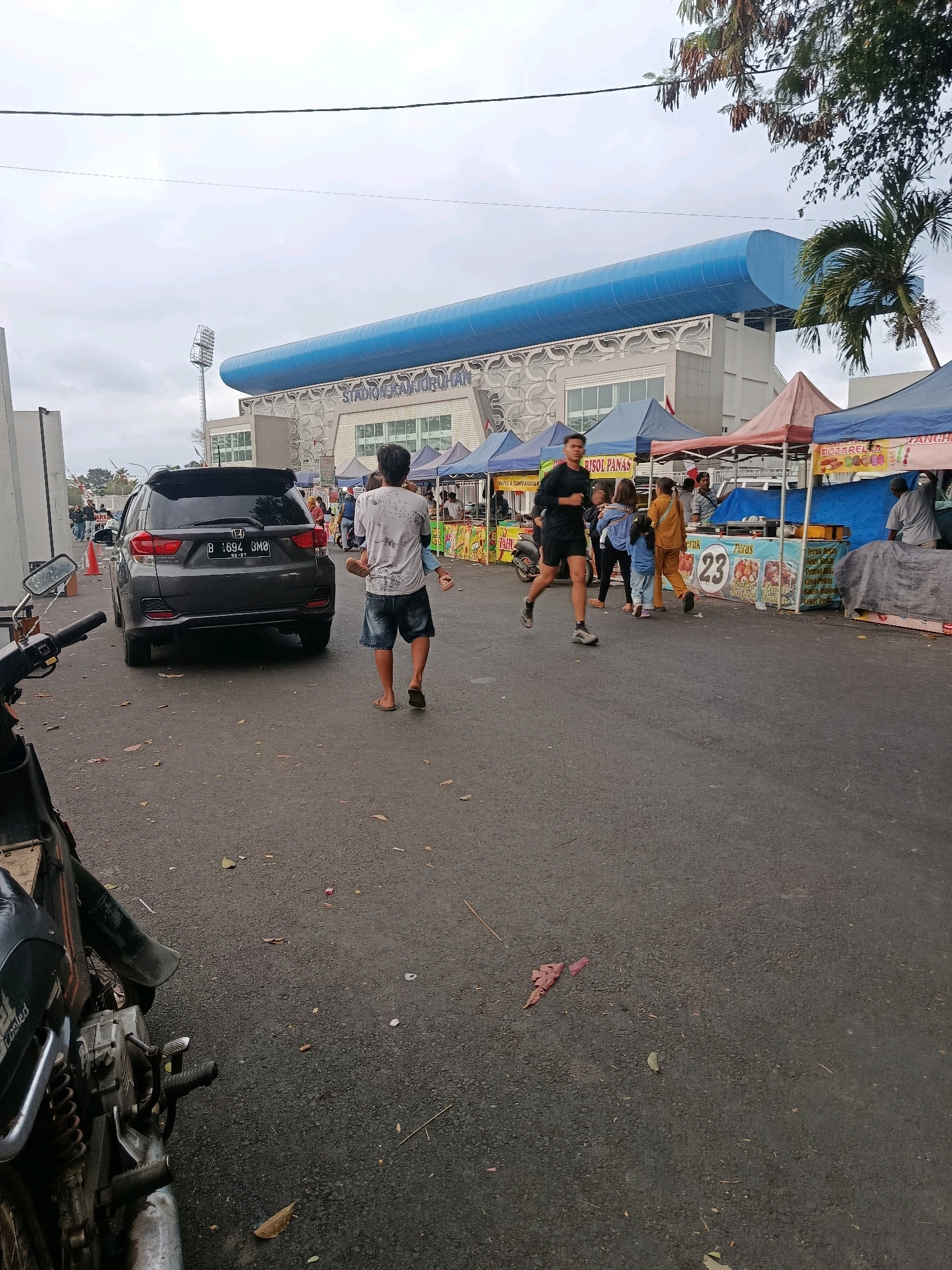 The image depicts a street scene in front of a stadium with a blue, curved roof. In the foreground, a dark-colored SUV is parked on the right side of the road. To the left of the SUV, a motorcycle is partially visible. A man wearing a white t-shirt and denim shorts is walking away from the camera, carrying a child in his left arm. To the right, a man in athletic wear is jogging. Several market stalls with colorful awnings are set up along the right side of the road, selling various food items. The ground is paved with asphalt and scattered with fallen leaves. The sky is overcast. The stadium's signage reads "STADION GELORA JURUHAY".