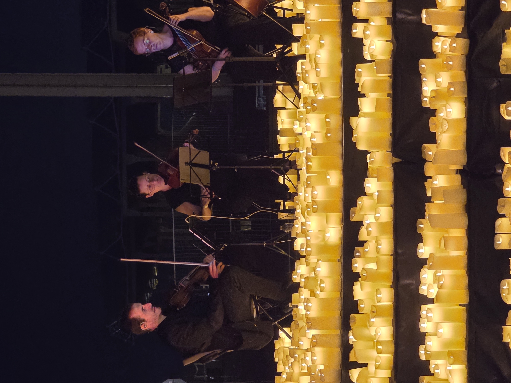 Three musicians are playing violins in front of a large display of illuminated candles at night. The musicians are positioned on a slightly raised platform, with the male musician on the left and two female musicians to his right. They are all dressed in dark clothing. The candles are arranged on tiered surfaces in the foreground, creating a warm, glowing atmosphere. The background is dark and appears to be an outdoor or semi-outdoor space with some industrial-looking structures visible. This scene appears to be a public event, possibly a concert or memorial, taking place in the city of Sant Adrià de Besòs, Spain.