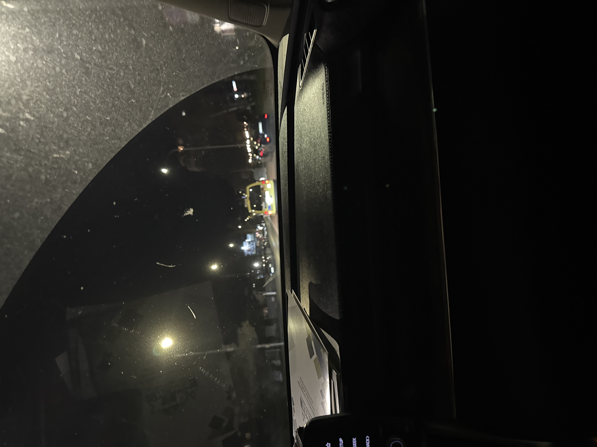 The image shows a view from inside a vehicle at night. The windshield is dirty and has a blurry view of city lights, possibly in Sofia, Bulgaria. There are streetlights, and the faint silhouettes of buildings visible through the glass. To the right of the windshield is the interior trim of the car, a dark-colored panel with what appears to be a passenger airbag label. At the bottom of the frame, there is a partially visible dashboard with some buttons. The overall scene is dark, illuminated by external light sources.