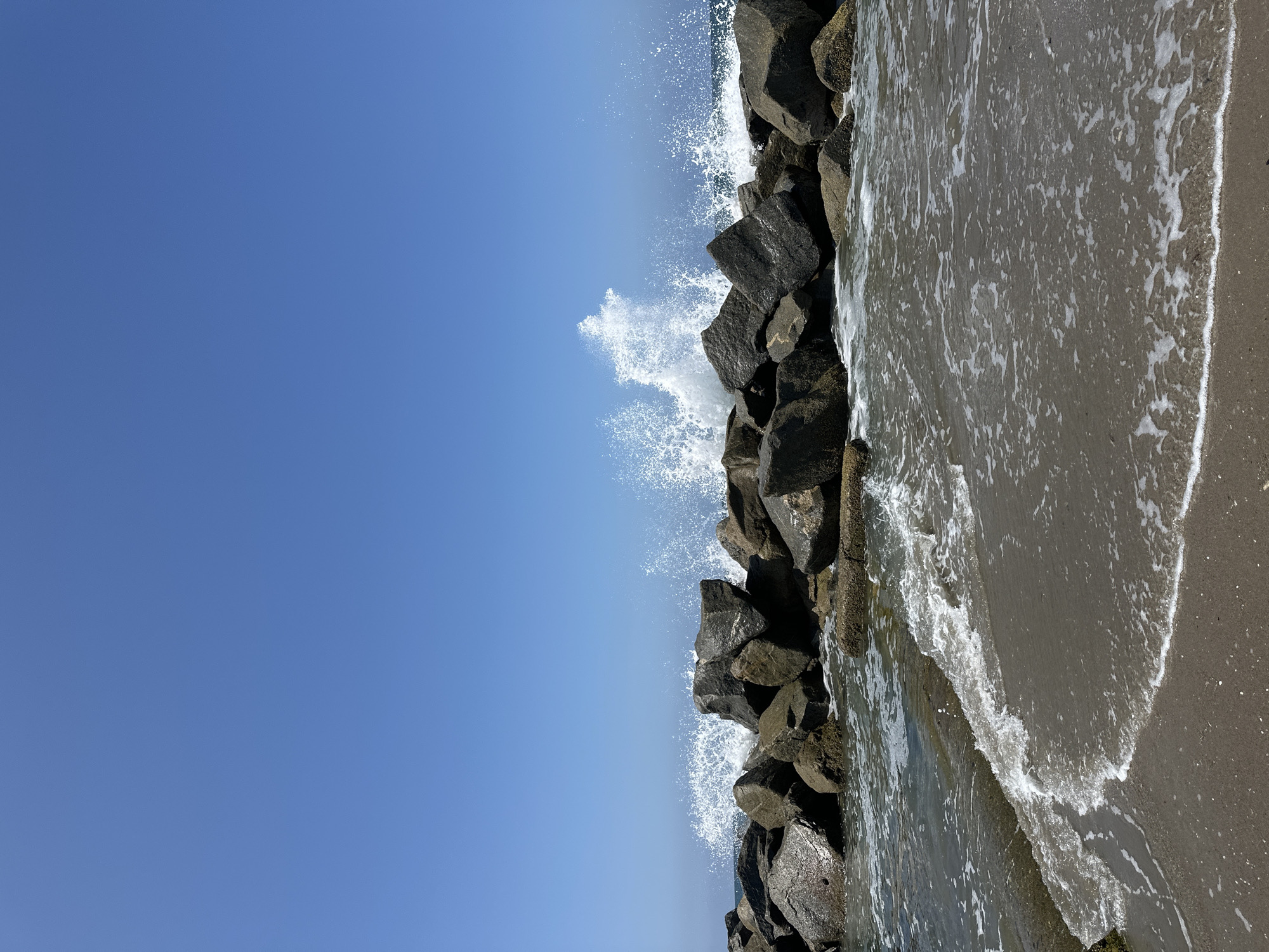 The image shows a seascape on a clear, sunny day. A breakwater made of large, dark rocks stretches across the middle ground, with waves crashing against them, sending white spray into the air. The ocean water is a deep blue, and a sandy beach with gentle waves lapping at the shore is visible in the foreground. The sky above is a clear, vibrant blue. This scene is characteristic of many coastal areas in the city of Los Angeles, United States of America, such as the breakwaters found at beaches like Santa Monica or Cabrillo Beach, which are designed to protect harbors and shorelines from wave action. There are no people or animals visible in the image.