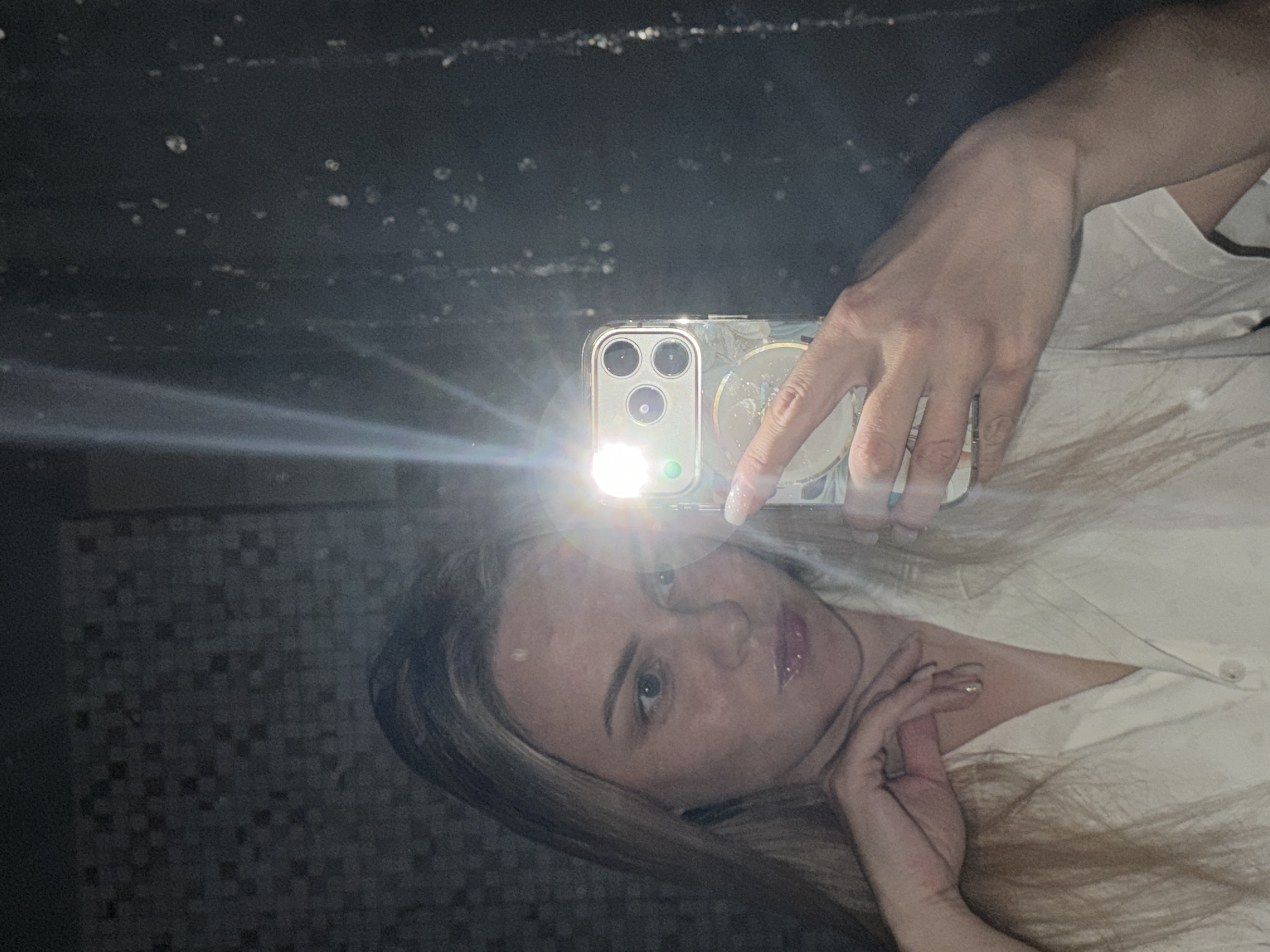 A woman is captured in a mirror selfie, holding a smartphone with its flash activated, causing a bright glare that partially obscures her face. She has light-colored hair, a neutral expression, and is looking directly at her reflection (and thus the camera). Her lips appear glossy. She is wearing a light-colored, possibly cream or white, collared shirt. Her fingernails are painted a pale shade.
The phone, which appears to be a modern iPhone model with multiple cameras, is held in a decorative or clear case. The bright flash illuminates the immediate area around the phone and her face.
The background reflected in the mirror is dark, revealing a checkered or tiled pattern in some areas, suggesting an indoor setting such as a bathroom or a room with distinctive wall tiles. The mirror surface itself shows some specks and streaks, possibly dust or moisture. The overall lighting condition appears dim, necessitated by the use of the phone's flash. No visible text is discernible in the image. The scene suggests a casual, personal moment captured in Votkinsk, Russia.