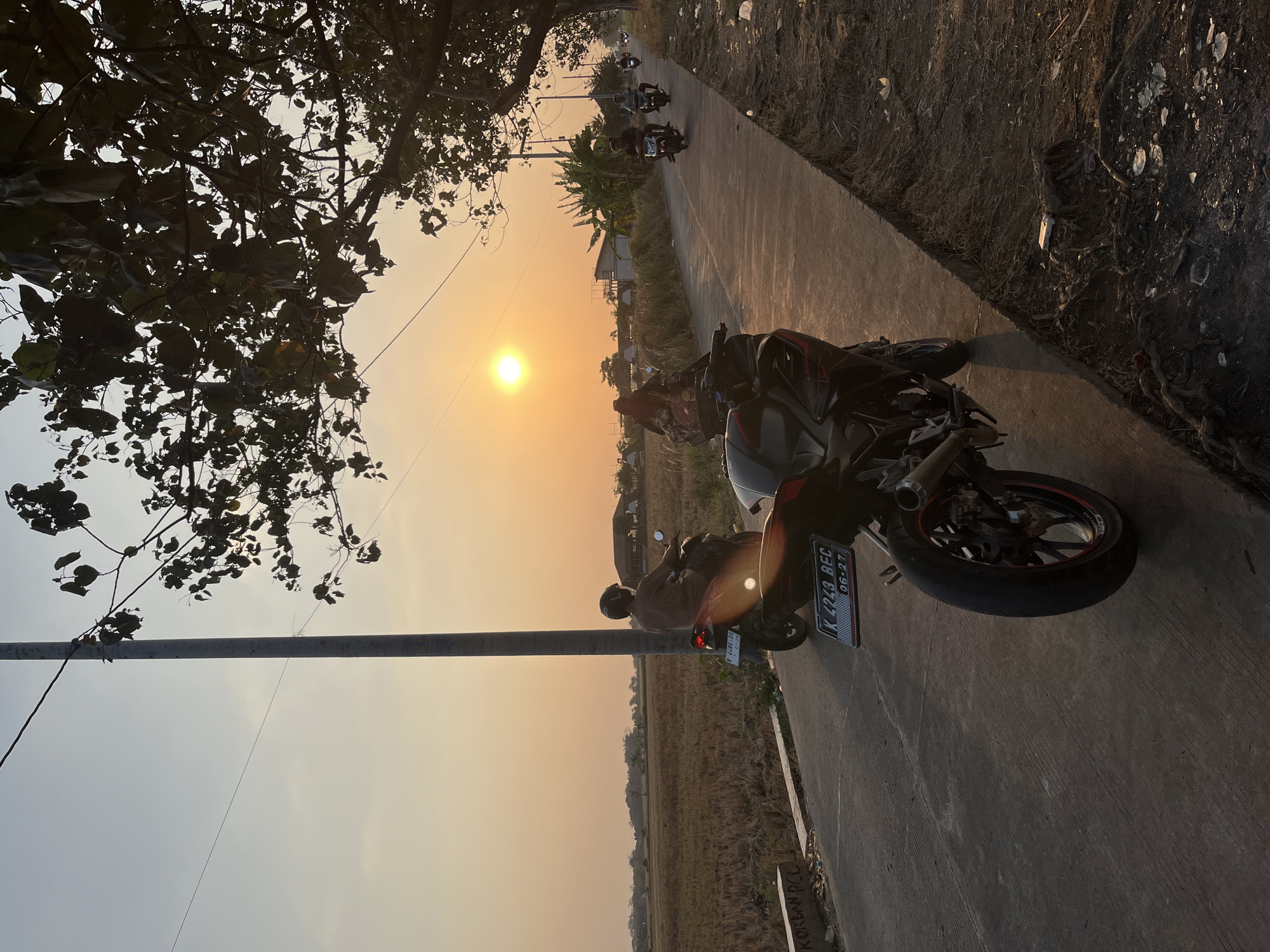 An outdoor, eye-level, vertical medium shot captures a road in Pecangaan, Indonesia, during sunset. The sun, a bright orange orb, hangs low in the sky, casting a warm glow over the scene. A road, made of concrete, stretches into the distance, flanked on the left by a dry, mowed field and on the right by a rough, dirt embankment covered in debris and roots. A single streetlight stands tall on the left side of the road, its shadow cast across the pavement.
In the foreground, a black motorcycle is parked on the right side of the road, its rear wheel and license plate clearly visible. The license plate reads "K 4243 BEC 06-27". Another motorcycle, ridden by a person wearing a brown shirt and helmet, is in the middle ground on the left side of the road, heading away from the camera. Further down the road, several other motorcycles, some with riders, are visible, all traveling in the same direction. In the background, there are houses and structures, and trees line the right side of the road, their branches silhouetted against the sunset. The overall atmosphere is tranquil and depicts a typical rural Indonesian scene at dusk.