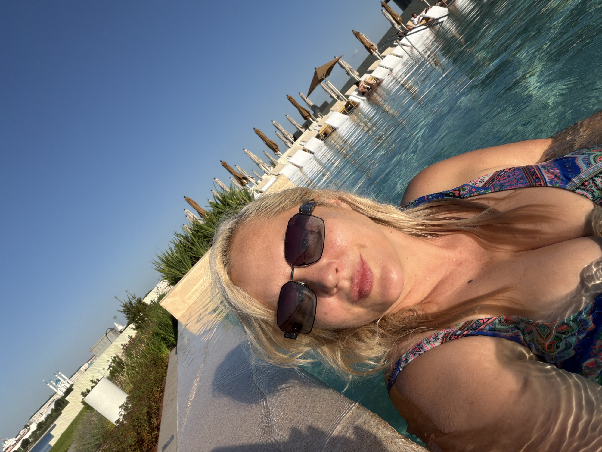 A woman with blonde hair and sunglasses is taking a selfie in a pool. She is wearing a colorful swimsuit. The pool has clear blue water and is surrounded by lounge chairs and umbrellas. In the background, there is a building complex with white walls and blue domes, possibly a hotel or resort. The sky is clear and blue.