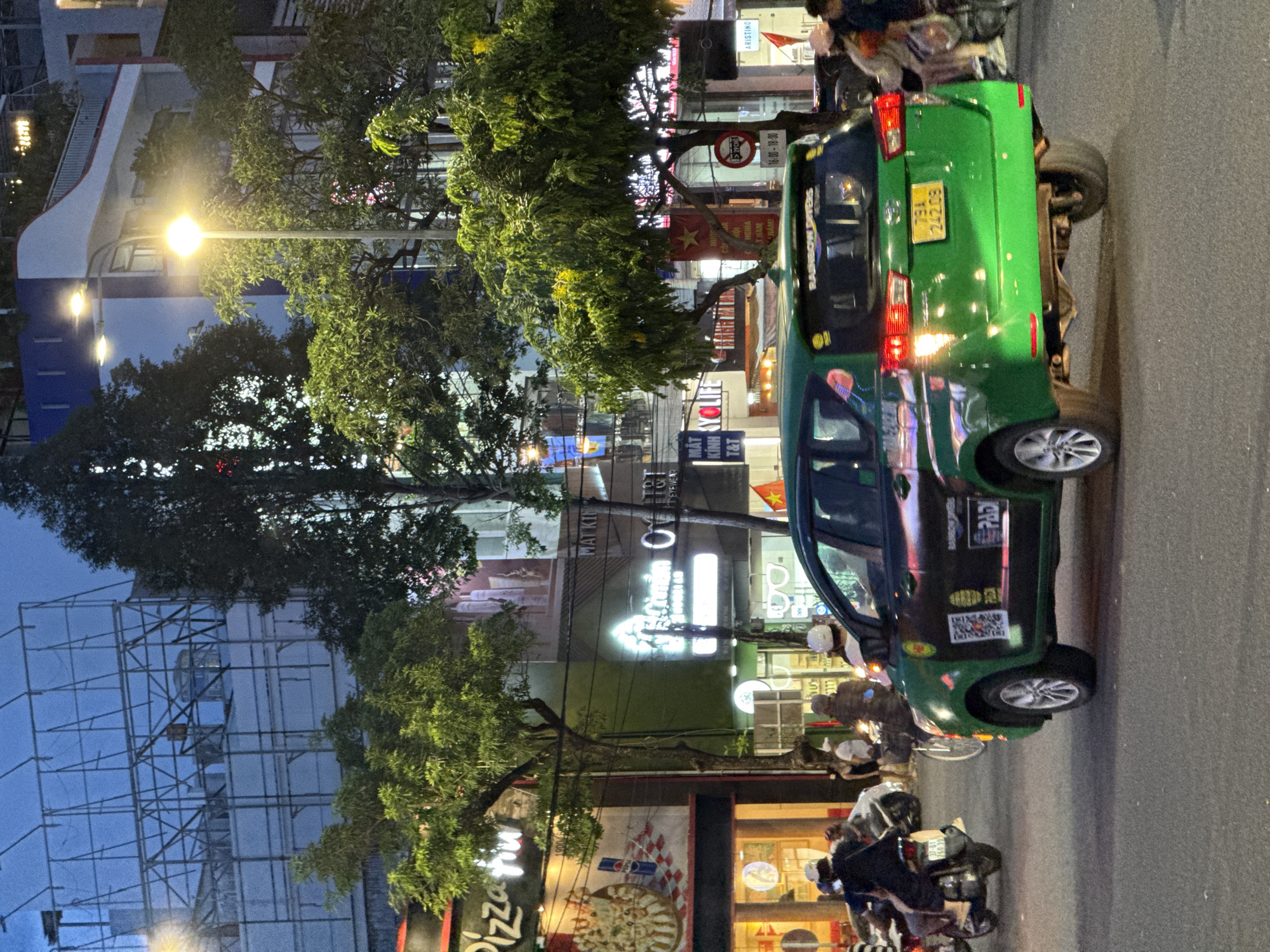 This image captures a street scene in Nha Trang, Vietnam, at dusk. A green taxi, possibly a Toyota Innova, is the central subject, with its rear visible to the viewer. The taxi has a license plate that reads "79A 242.09". Advertisements and decals are visible on the side of the taxi, including a QR code and text that says "RAINBOW DIVERS". Other vehicles, including motorcycles and bicycles, are present on the road, indicating traffic. Several people are seen on motorcycles and sidewalks, some wearing helmets. In the background, buildings with illuminated signs line the street. One sign appears to be for "Pizza Hut". Trees with lush green foliage are interspersed with the buildings, and streetlights illuminate the scene. The overall atmosphere suggests a bustling urban environment.