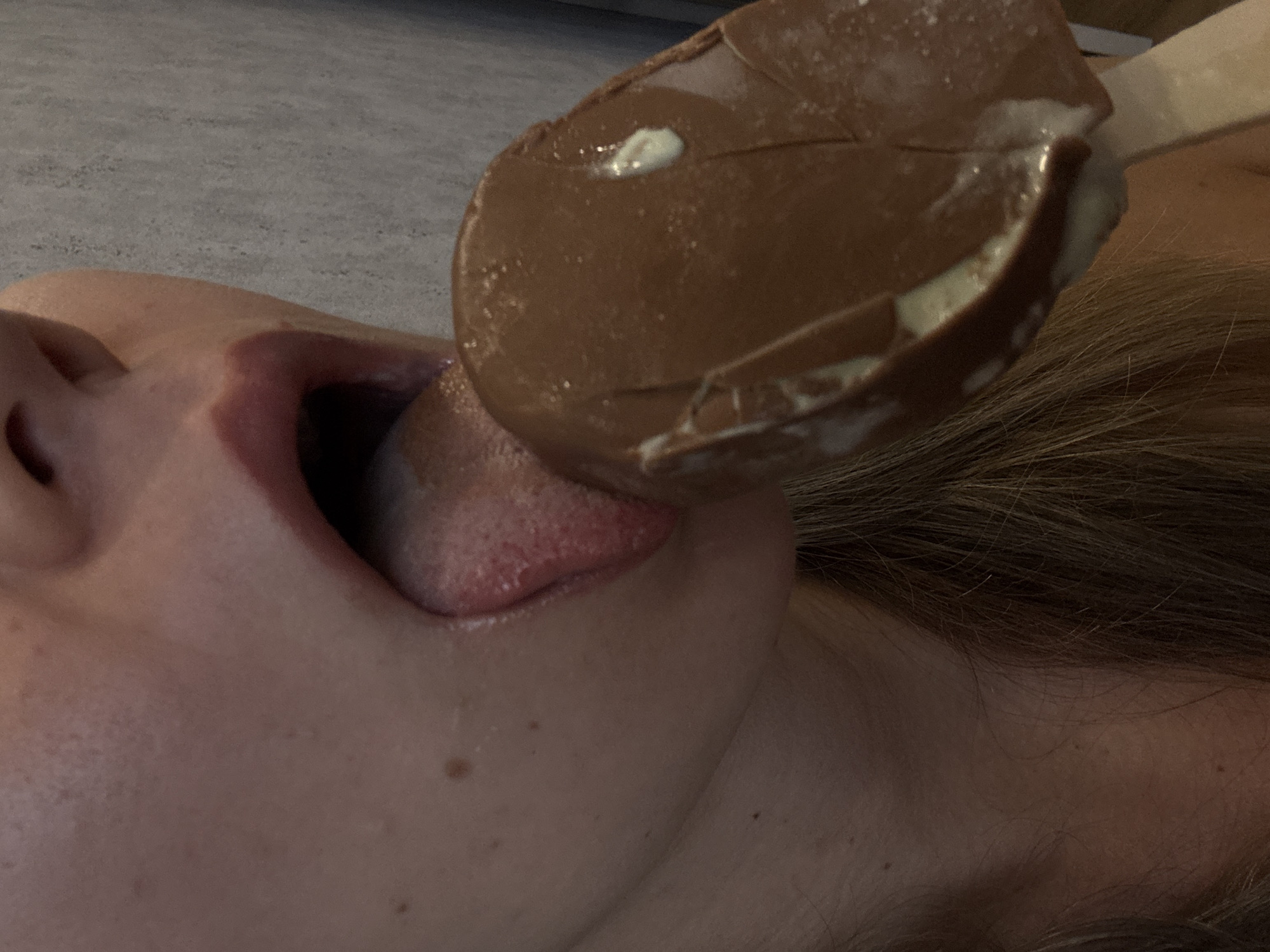 The image is a close-up shot of a person's mouth and chin as they lick a chocolate-covered ice cream bar. The person's lips are slightly parted, and their tongue is visible, reaching out to taste the ice cream. The ice cream bar is mostly covered in chocolate, with some white creamy filling visible. The background appears to be a textured surface, possibly a table or countertop, in a neutral gray tone. The lighting is soft, casting subtle shadows and highlighting the textures of the skin and the ice cream. The focus is sharp on the mouth and ice cream, creating a sense of intimacy and sensory experience. There is no visible text in the image. The overall impression is one of enjoying a sweet treat.