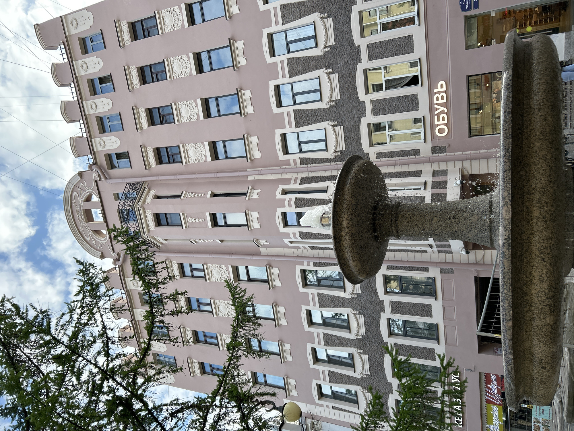 The image shows a building in Saint Petersburg, Russia, as evidenced by Cyrillic text and architectural style. The building is multiple stories tall with a pink facade, decorative window frames, and a rounded corner featuring a balcony. A stone fountain is in the foreground, with water visible in motion. A tree with green foliage partially obscures the view of the building's lower section. The sky is visible in the background, with clouds suggesting a daytime scene. The word "Обувь" (Shoes) is seen on the building, indicating a shoe store. Other signs are also present but difficult to discern. No people are directly visible.