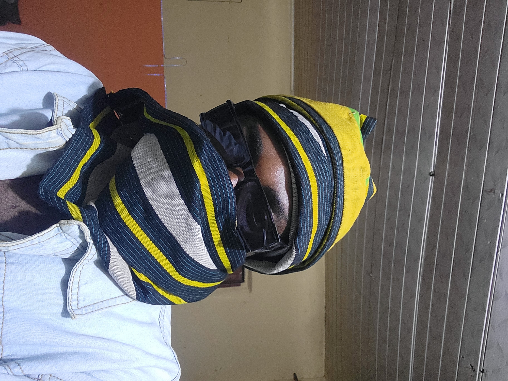 An image, likely taken in Jos, Nigeria, shows a close-up, rotated portrait of a person. The individual, who appears to have a dark skin tone, is seen from the side, with the right ear, part of the cheek, and forehead visible. They are wearing dark-rimmed glasses. Their head is adorned with a distinctive head covering, possibly a cap or wrapped fabric, featuring bold, alternating stripes of black (or very dark blue), bright yellow, and white.
On the left side of the frame, the shoulder and upper back of a light blue garment, possibly denim or a similar fabric with visible stitching, can be seen. The person's posture suggests they might be resting or looking downwards.
The background is composed of several distinct sections. In the upper left, an orange-brown wall or surface is visible, against which a faint wire object, possibly a hanger, can be discerned. This transitions to a plain beige wall in the central background. To the right, a dark gray, vertically ridged surface, resembling corrugated metal or paneling with faint white lines, occupies a significant portion of the background. The lighting is consistent with an indoor setting, casting some shadows. No visible text is present in the image.