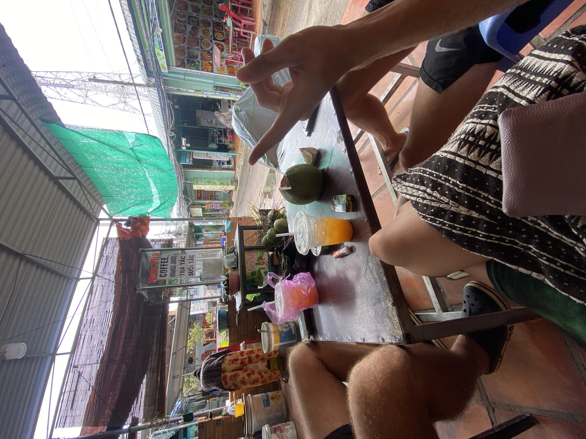 The scene captures a casual outdoor setting in Phu Thuy Ward, Vietnam, likely a street-side cafe or eatery. A group of people are seated around a weathered metal table, enjoying drinks and snacks. On the table are coconuts, beverages in plastic cups and bags, and other items. People are seen in the foreground and background, some identifiable by their attire, such as shorts, patterned clothing, and footwear. The surrounding environment includes street-side architecture, signage, overhead roofing, and typical urban elements of Phu Thuy Ward.
