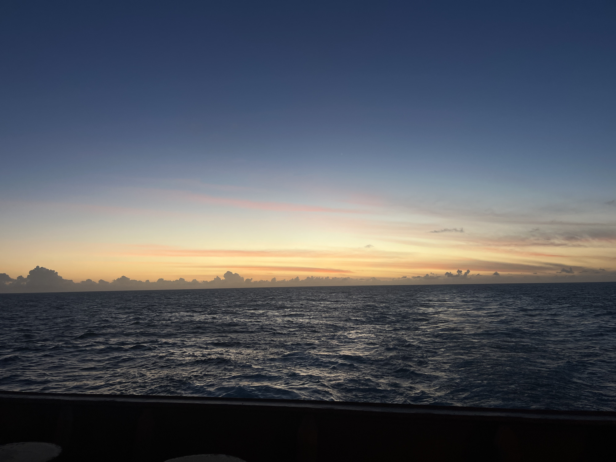 The image depicts an ocean view at dusk or dawn. The sky transitions from a dark blue at the top to a light orange and pink near the horizon. There are a few scattered clouds near the horizon. The ocean is dark and wavy. A dark structure, possibly a boat railing, is visible in the foreground. The location is on the ocean, the city and country are unknown.