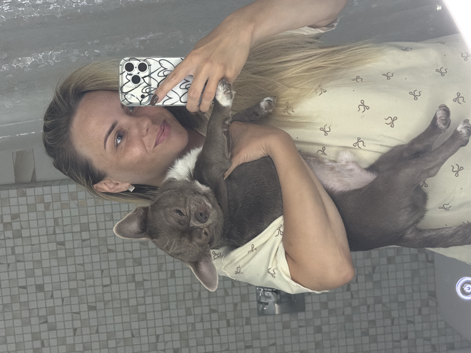 A young woman is taking a selfie in a bathroom mirror, holding a small grey dog. She has blonde hair and is wearing a light-colored shirt with a bow pattern. The dog is looking down and appears relaxed. The bathroom has tiled walls and a shower curtain.