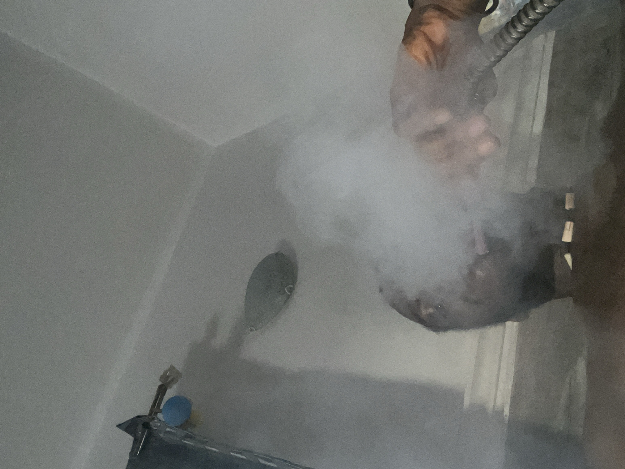 The image depicts a steamy bathroom environment. A showerhead is visible on the wall, and a hand holding a hose is positioned in the upper right corner. The steam obscures many details.