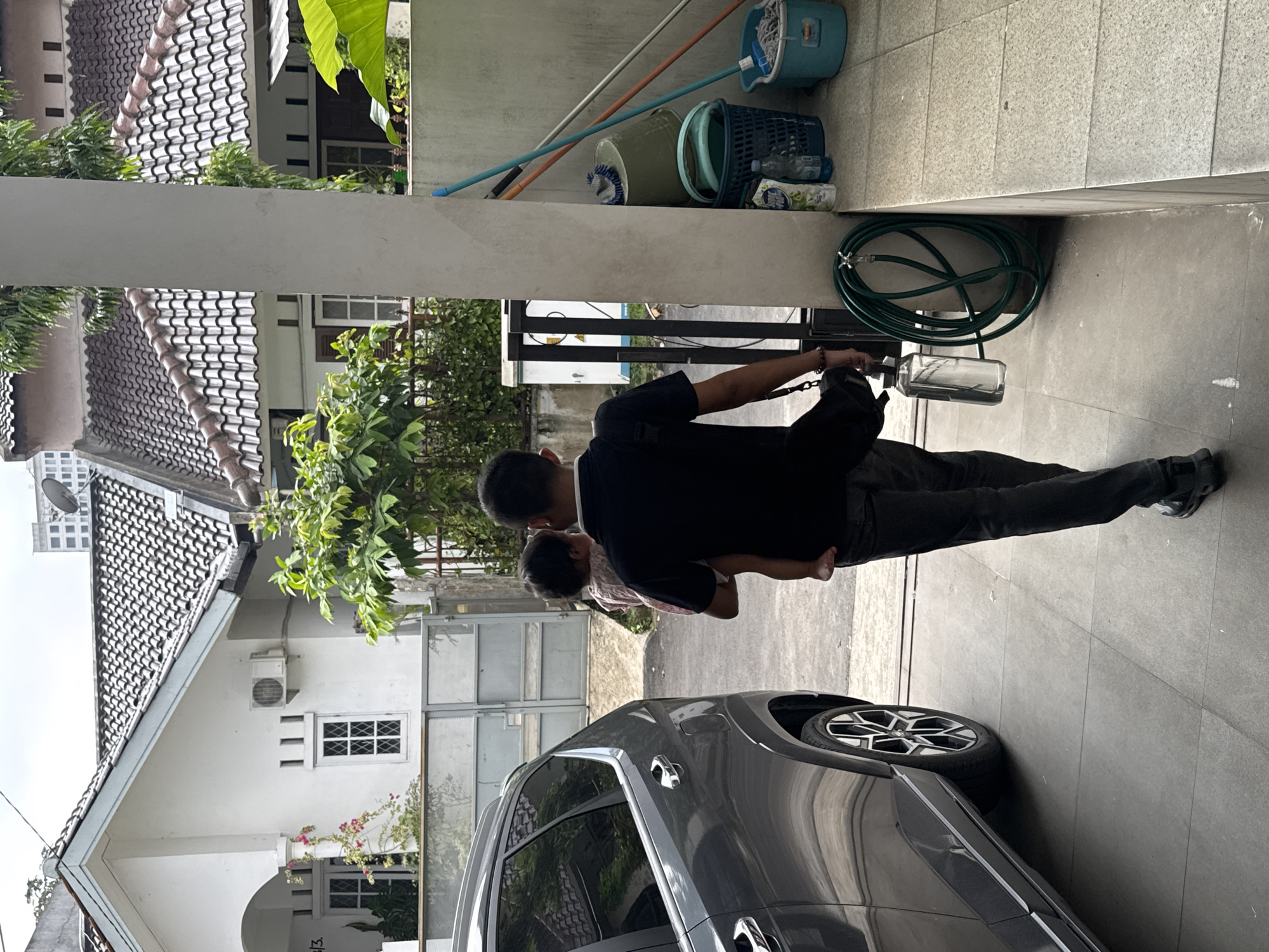 A man in a black shirt and dark pants holds a child in his arms while walking in a driveway. The scene occurs in Bekasi, Indonesia, as evidenced by the architecture and general surroundings. A grey car is parked on the driveway. A coiled green garden hose hangs on the wall. Cleaning supplies, including buckets and mop, are on a ledge. Houses with tiled roofs are visible in the background. The driveway is paved with grey tiles. The man is holding a clear container of liquid.
