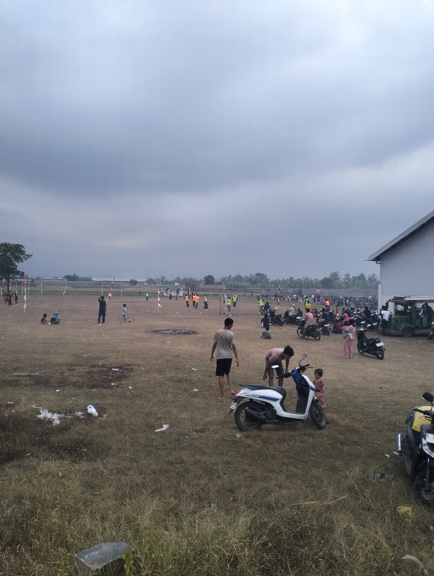 The image depicts an outdoor scene, possibly a field or park, in Sokaraja, Indonesia, under a cloudy sky. The ground is covered in dry grass and scattered with white plastic trash. In the mid-ground, a group of people are engaged in various activities. Some are standing and conversing, while others are tending to motorcycles parked in clusters.
In the foreground, there are two scooters. One is white and blue, and the other is black with a yellow seat. A young child in a pink dress stands near the white and blue scooter. Further back, a man wearing a light brown t-shirt and dark shorts is standing. A group of people are visible in the far background, some appearing to be participating in a sporting event or gathering. Flags on poles are also present in the distance, suggesting a possible local event or celebration. A building with a white wall and a grey roof is situated on the right side of the image. The overall impression is one of a casual gathering or event taking place in a somewhat unkempt outdoor space.