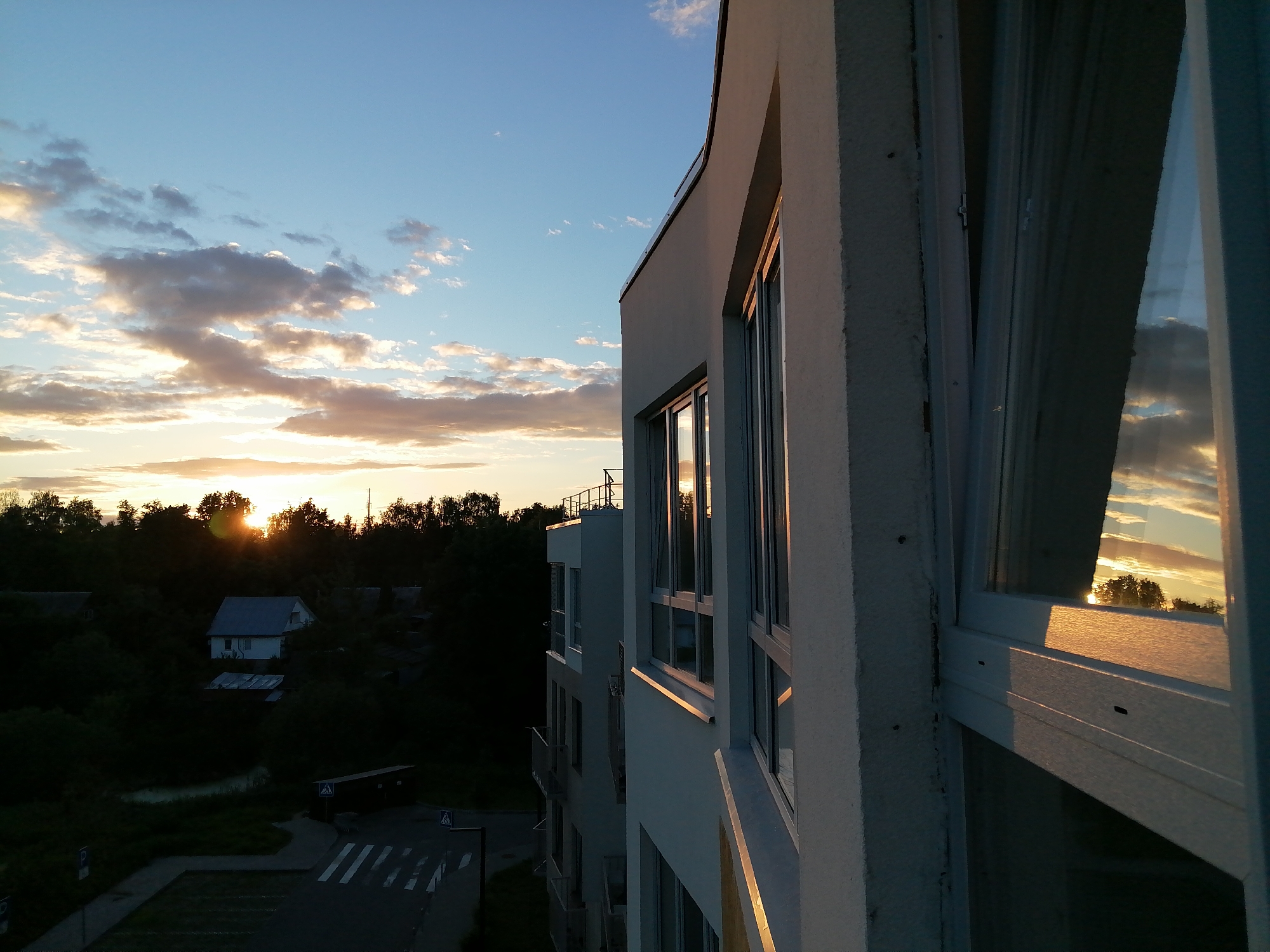 The image depicts a modern apartment building with large windows reflecting the sunset. The building's exterior is a light, textured white. The windows are framed in white, and through them, the warm hues of the setting sun are visible, along with scattered clouds. Below the building, a dark tree line is silhouetted against the sky. The lower portion of the image shows a street with a pedestrian crossing, some parked cars, and a few low-lying buildings partially obscured by darkness. The scene is set outdoors in what appears to be a suburban or semi-urban environment during the golden hour of sunset.