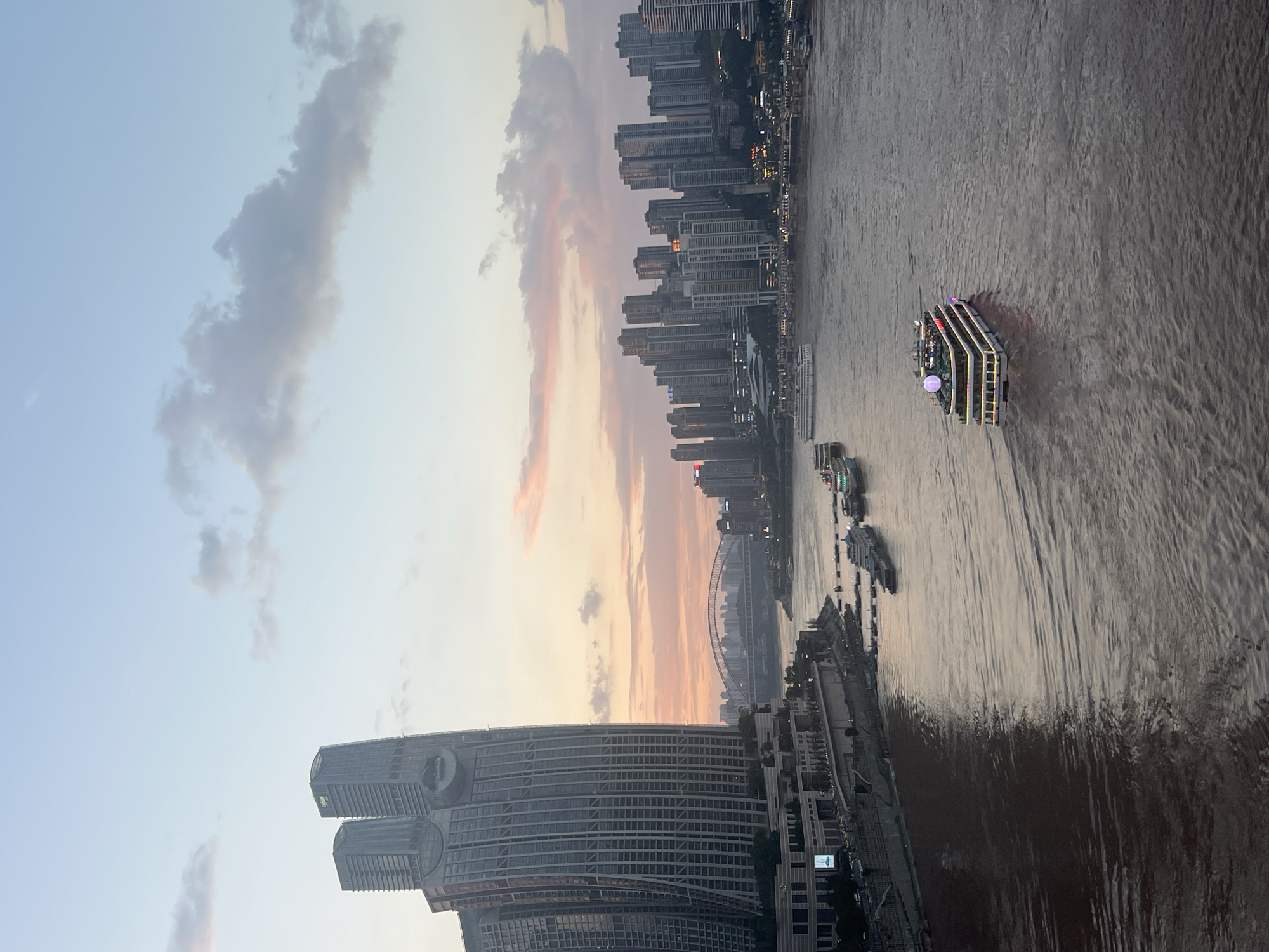 This is a long-shot, wide-angle, eye-level, slightly elevated, vertical photograph of a wide, brown river at dusk, with a city skyline and a bridge in the background. The scene is located in Nan'an District, China. On the left, a modern, tall, glass-facade building dominates the foreground. In the middle ground, the river is occupied by several boats, including a large tour boat with illuminated decks, moving from right to left. The riverbanks are lined with buildings, some of which are lit. On the far side of the river, a dense collection of high-rise buildings forms the city skyline, and a long, arched bridge spans the river in the distance. The sky above is a gradient of pale blue and soft pinks, with scattered clouds, indicative of sunset. The overall atmosphere is calm and urban.