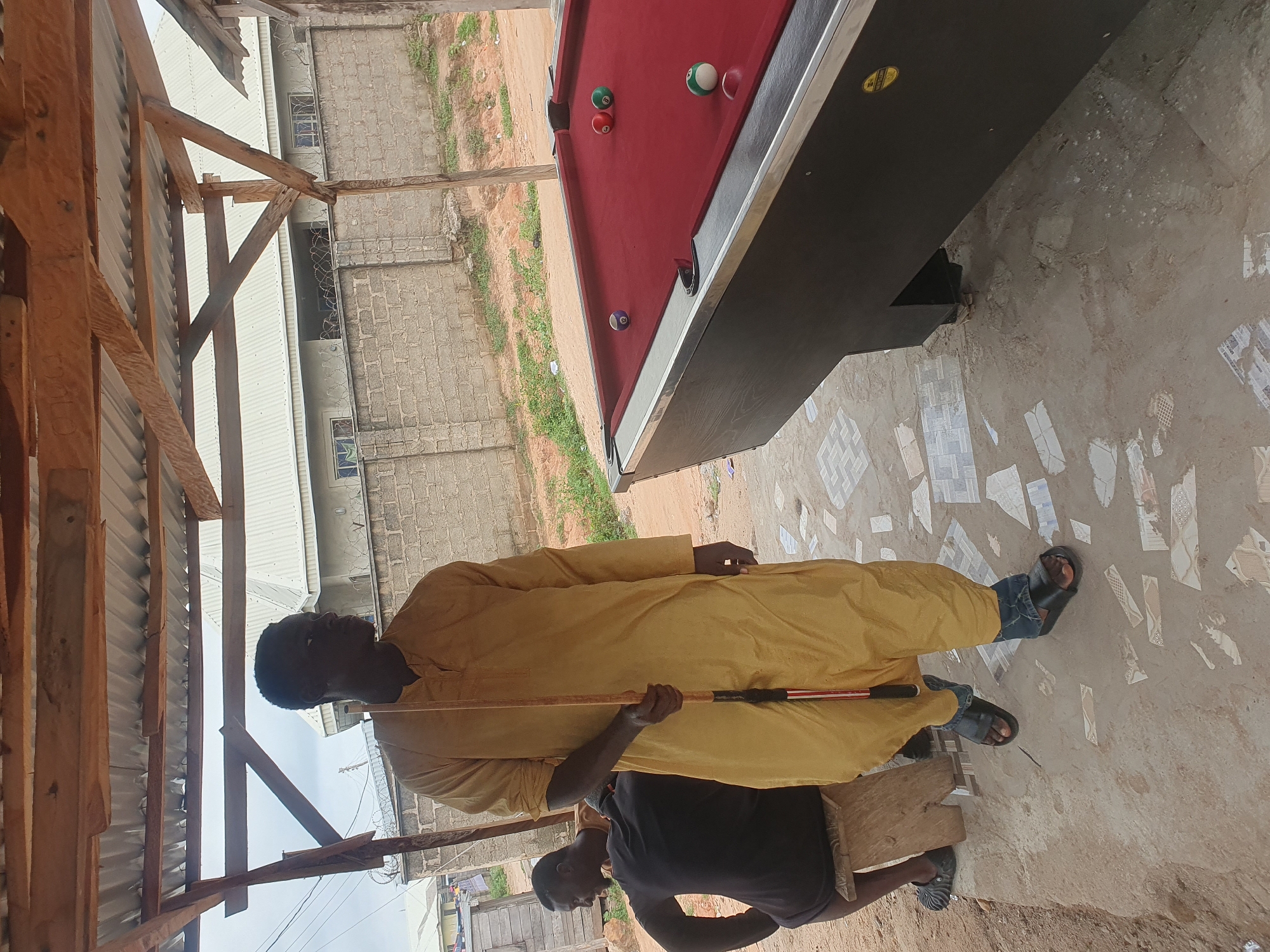 In Lokoja, Nigeria, a man in a yellow kaftan stands outdoors next to a pool table. He holds a pool cue vertically. To his left, another man wearing a black t-shirt is seated on a wooden stool. A red felt pool table with several balls is visible to the right of the main subject. The environment appears to be an informal outdoor setting with a corrugated metal roof supported by wooden beams. In the background, a brick wall with barbed wire separates the area from a building with white roofing and windows. The ground is a mixture of dirt and scattered tile fragments.