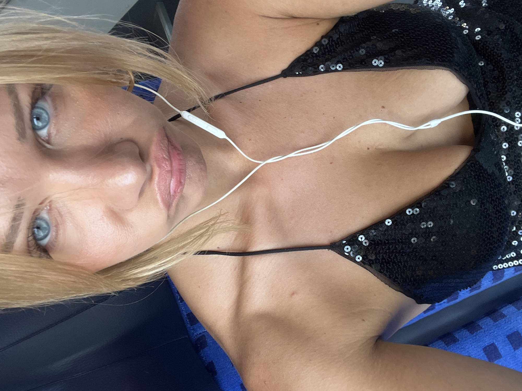 A close-up, selfie-style portrait features a blonde woman with striking blue eyes and pouty lips, wearing a black sequined dress and white earbuds. She is seated, indicated by the blue fabric with a checkered pattern visible in the background, likely a seat in a mode of public transportation or a waiting area. The lighting suggests an indoor or shaded outdoor setting. The woman's expression is ambiguous, possibly playful or slightly annoyed. The overall tone of the image is casual and personal. There is no discernible connection to Mühldorf a. Inn, Germany, as no elements in the image suggest a specific location within that city.