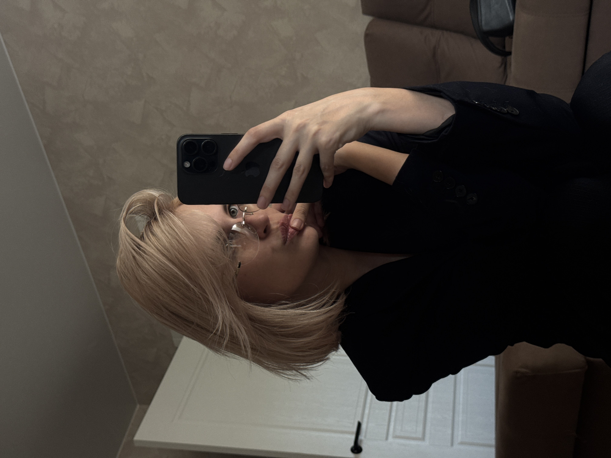 A young woman with blonde hair is taking a selfie using a black smartphone. She is wearing glasses and a black blazer. The background includes a beige wall, a white door with a black handle, and part of a brown sofa. The location is an interior space. The precise location in Saratov, Russia, cannot be determined from the image.