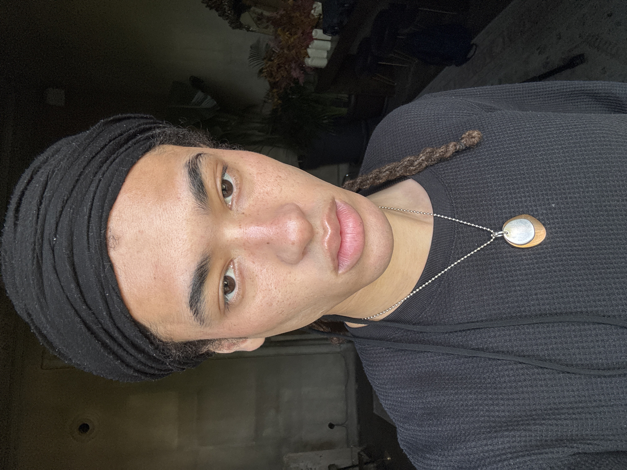 The image is a close-up, chest-up portrait of a person, originally rotated 90 degrees clockwise. The individual has medium-dark skin, dark brown eyes, and full, slightly glossy lips with visible freckles across their nose and cheeks. Their head is partially covered by a black head wrap or turban, and a brown dreadlock is visible extending over their right shoulder and chest.
They are wearing a black, waffle-knit long-sleeved shirt with a black drawstring across the chest. A silver bead chain necklace adorned with a unique two-toned pendant, comprising both silver and a warm-toned material (possibly copper or wood), hangs around their neck. The person looks directly at the camera with a neutral and composed expression.
The background is dark, out of focus, and appears to be an indoor setting with subdued lighting. Vague shapes that might be dried plants or decor are discernible in the upper left, suggesting an interior space. There is no specific activity depicted other than posing for the photograph. The time of day and weather are not evident. There is no visible text in the image. The location context provided is Madrid, Spain.