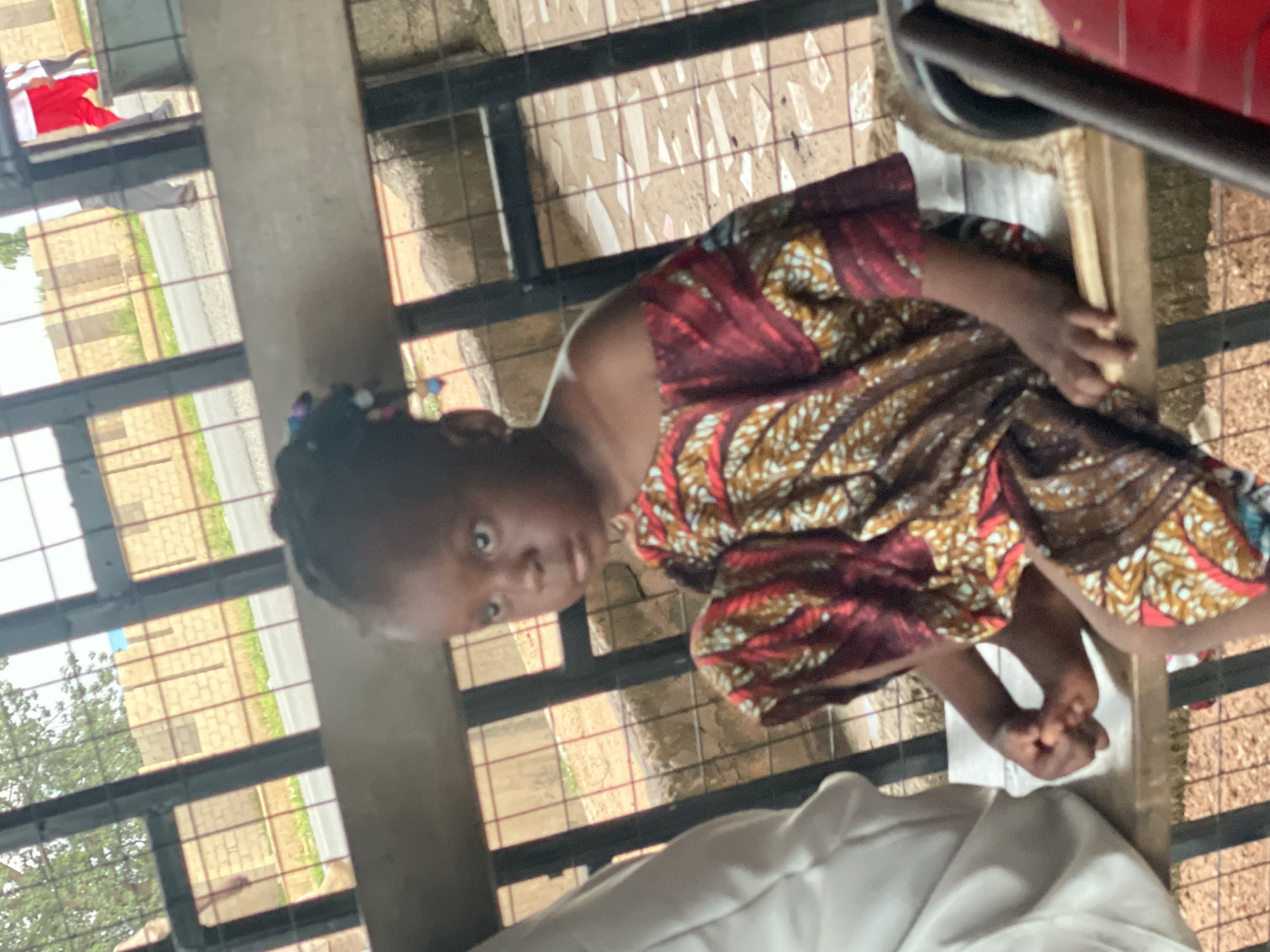 A young Black girl is seated on a wooden bench. She is wearing a colorful, off-the-shoulder dress with a geometric pattern in red, brown, and yellow. Her hair is braided and adorned with colorful beads. She is looking to her left with a pensive expression. To her left, a person in a white garment is partially visible. The bench is situated outdoors, with a black metal fence in the background. Beyond the fence, construction work is evident with unfinished brick walls and scattered debris. A paved road is also visible. The overall setting appears to be an outdoor area in Jalingo, Nigeria, possibly near a construction site or a public gathering space.