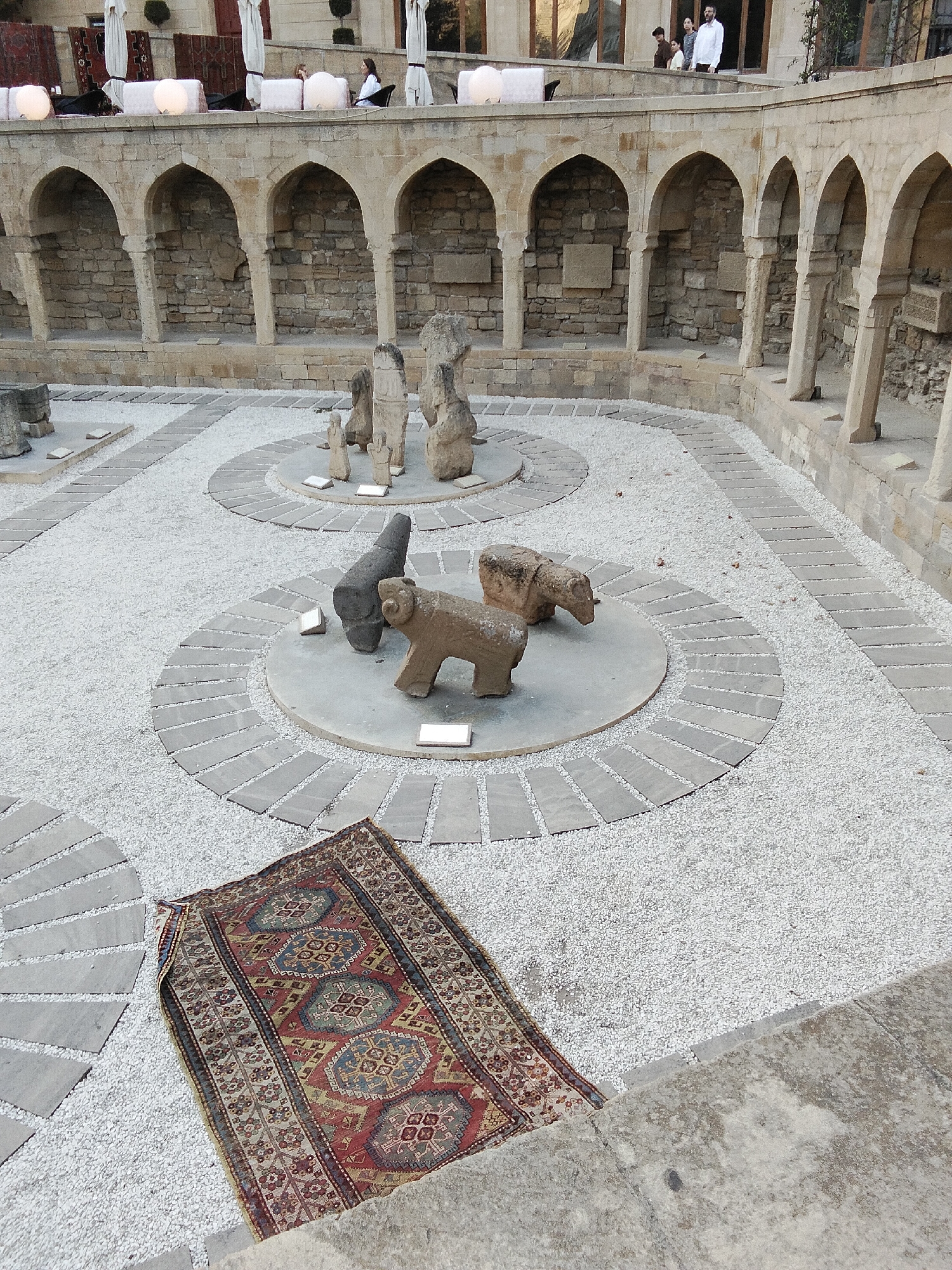 The image depicts an outdoor courtyard with a stone arcade in the background. The courtyard is paved with white gravel and features several circular stone platforms with ancient stone sculptures. One platform displays a sculpture of a ram and another dark, abstract-shaped sculpture. Further in the background, a group of taller, slender stone figures are arranged on a circular platform. A folded, ornate rug is placed on the gravel in the foreground, partially on a stone pathway. In the upper background, an elevated walkway with people seated at tables and umbrellas suggests a cafe or restaurant area. The architecture and sculptures suggest a historical or archaeological site. The location is identified as being in the Sabail Raion, Azerbaijan.